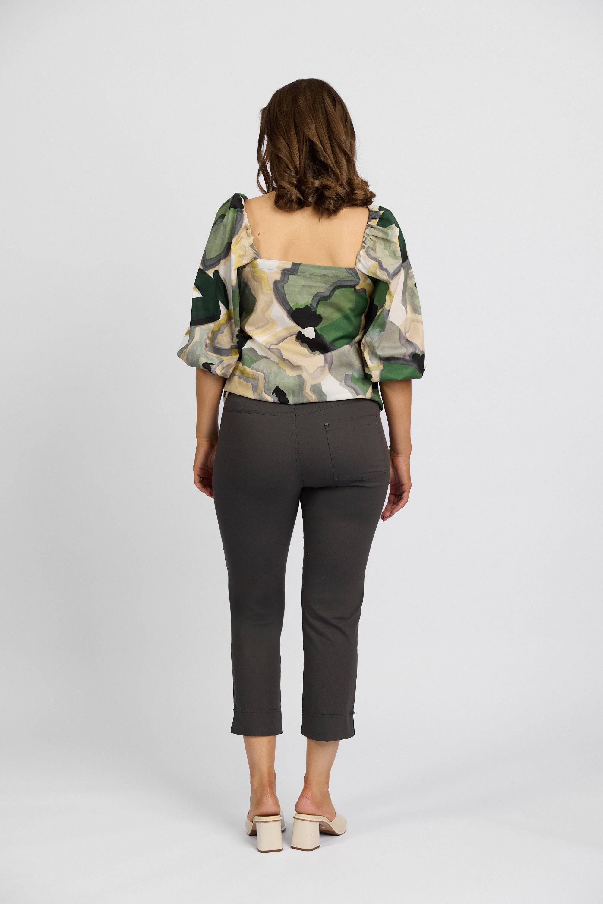 A woman stands facing away, wearing a green and beige patterned puff-sleeve blouse, Vassalli Paris Tour Capri - Beluga dark gray cropped pants, and light heeled sandals against a white background.
