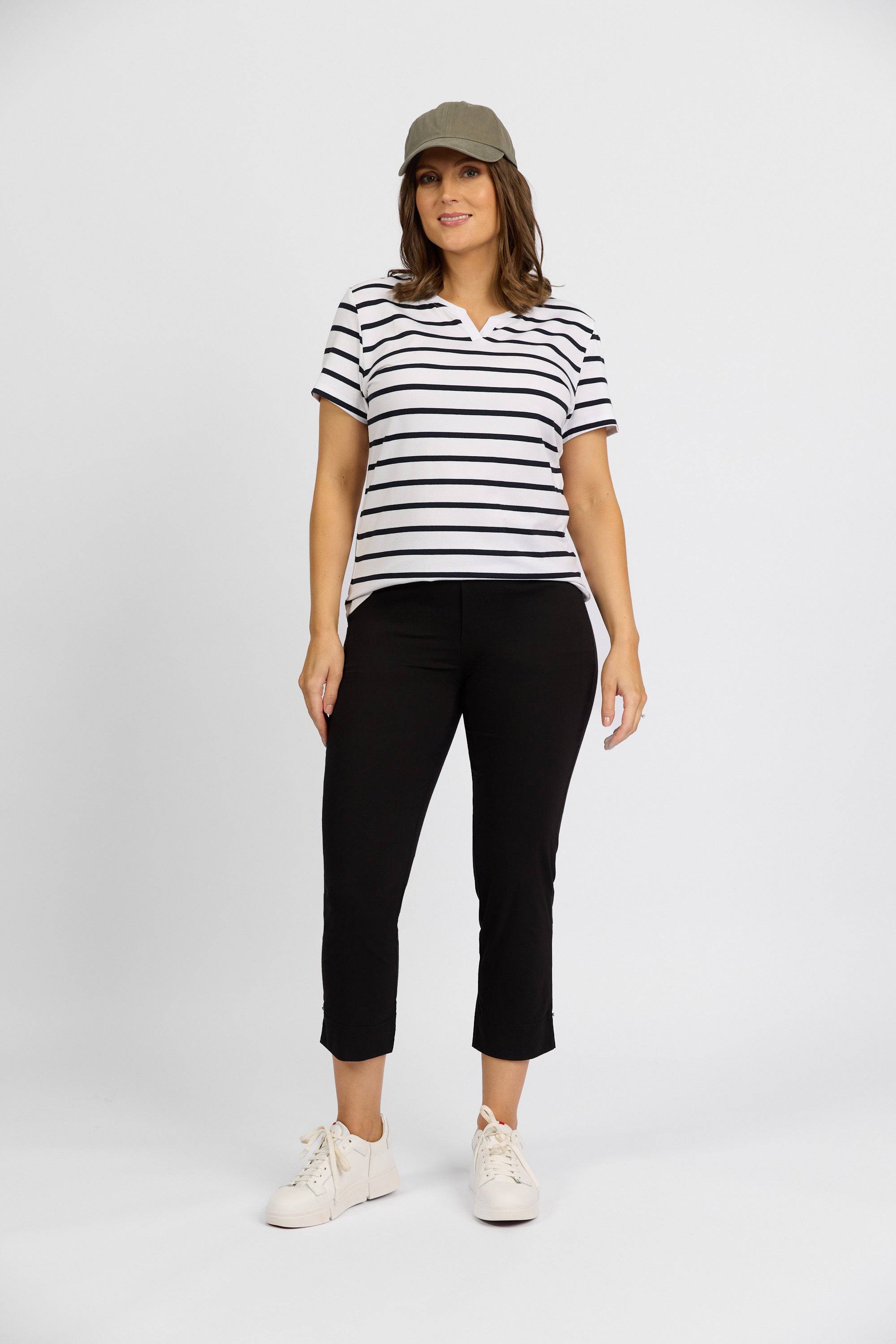 A woman stands against a white background wearing a green cap, black-and-white striped short-sleeve top, Vassalli Paris Tour Capri - Black pants, and white sneakers. She faces forward with a relaxed posture and a slight smile.