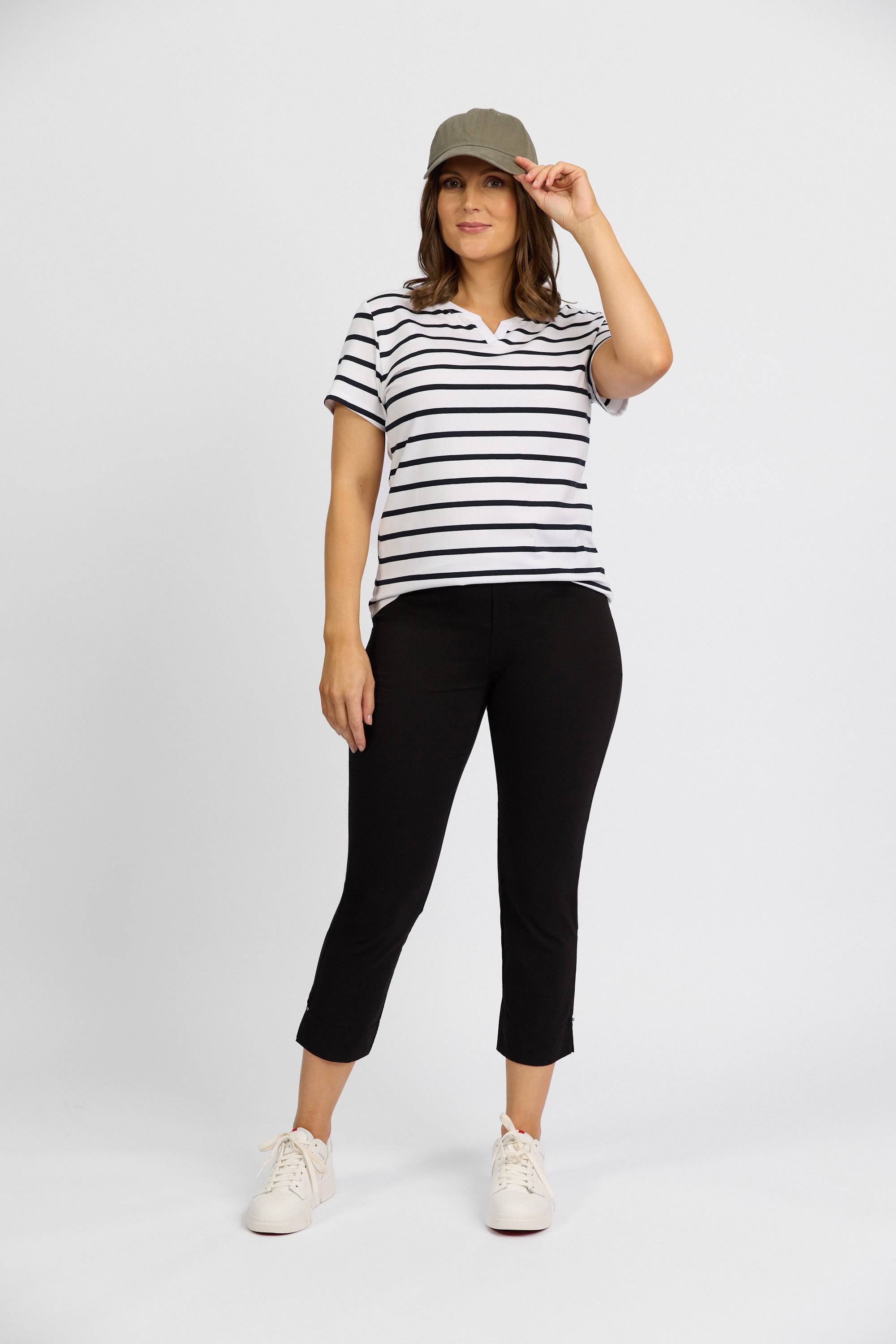 A woman stands against a white background, smiling and holding her cap as she models Vassalli’s Paris Tour Capri - Black pants, paired with a gray cap, a white and black striped short-sleeve shirt, and white sneakers.