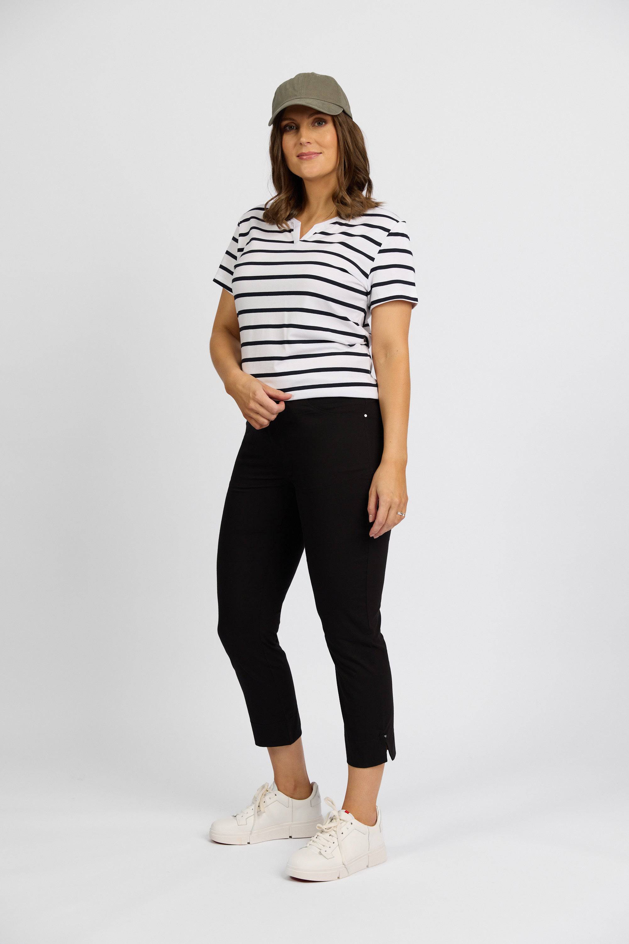 A woman in a green cap, Vassalli short-sleeve white shirt with black stripes, Paris Tour Capri - Black pants by Vassalli, and white sneakers stands against a plain white background, facing the camera and smiling slightly.