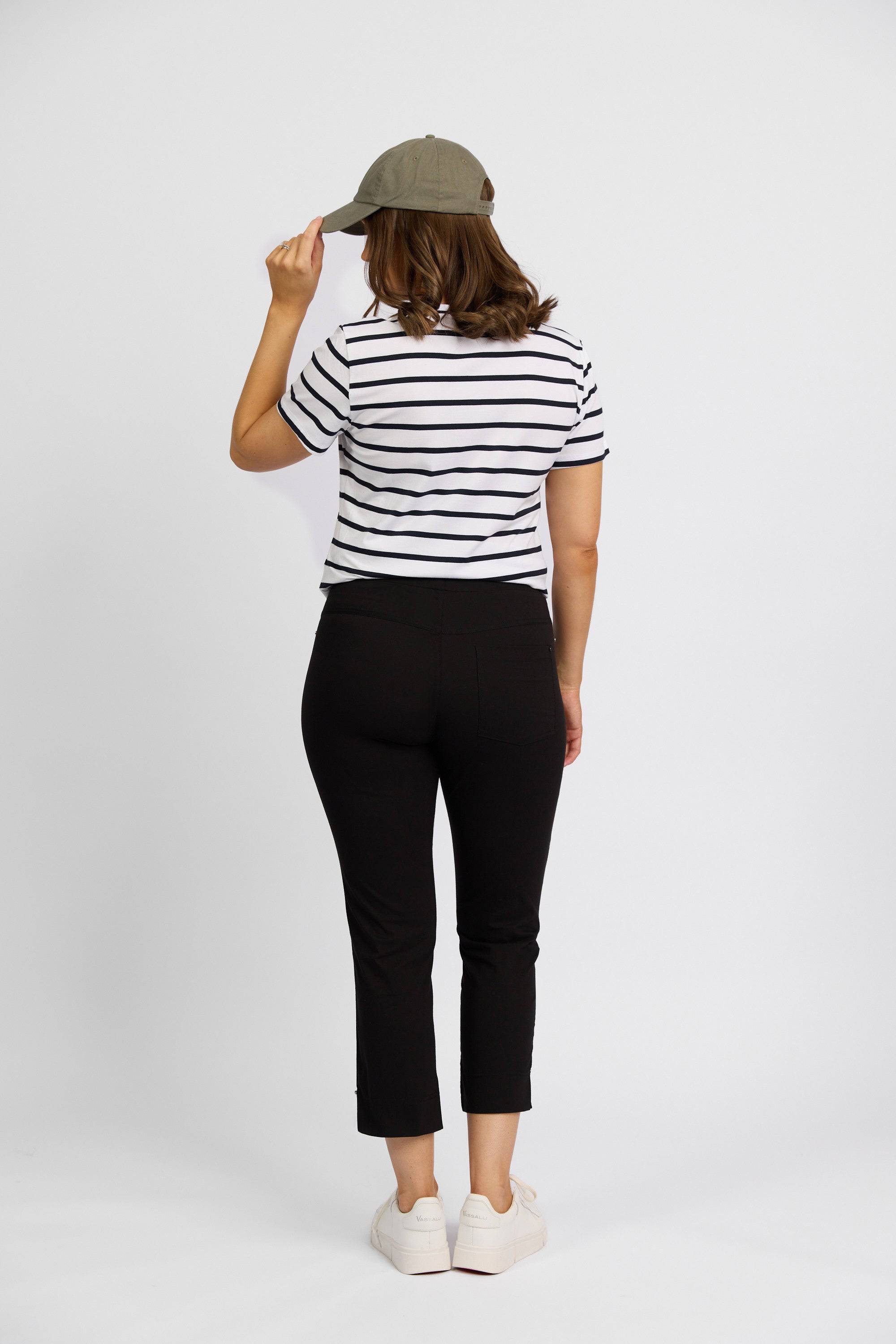 A person faces away, wearing a Vassalli striped short-sleeve shirt, Paris Tour Capri - Black by Vassalli, white sneakers, and holding a green cap. The background is plain white.