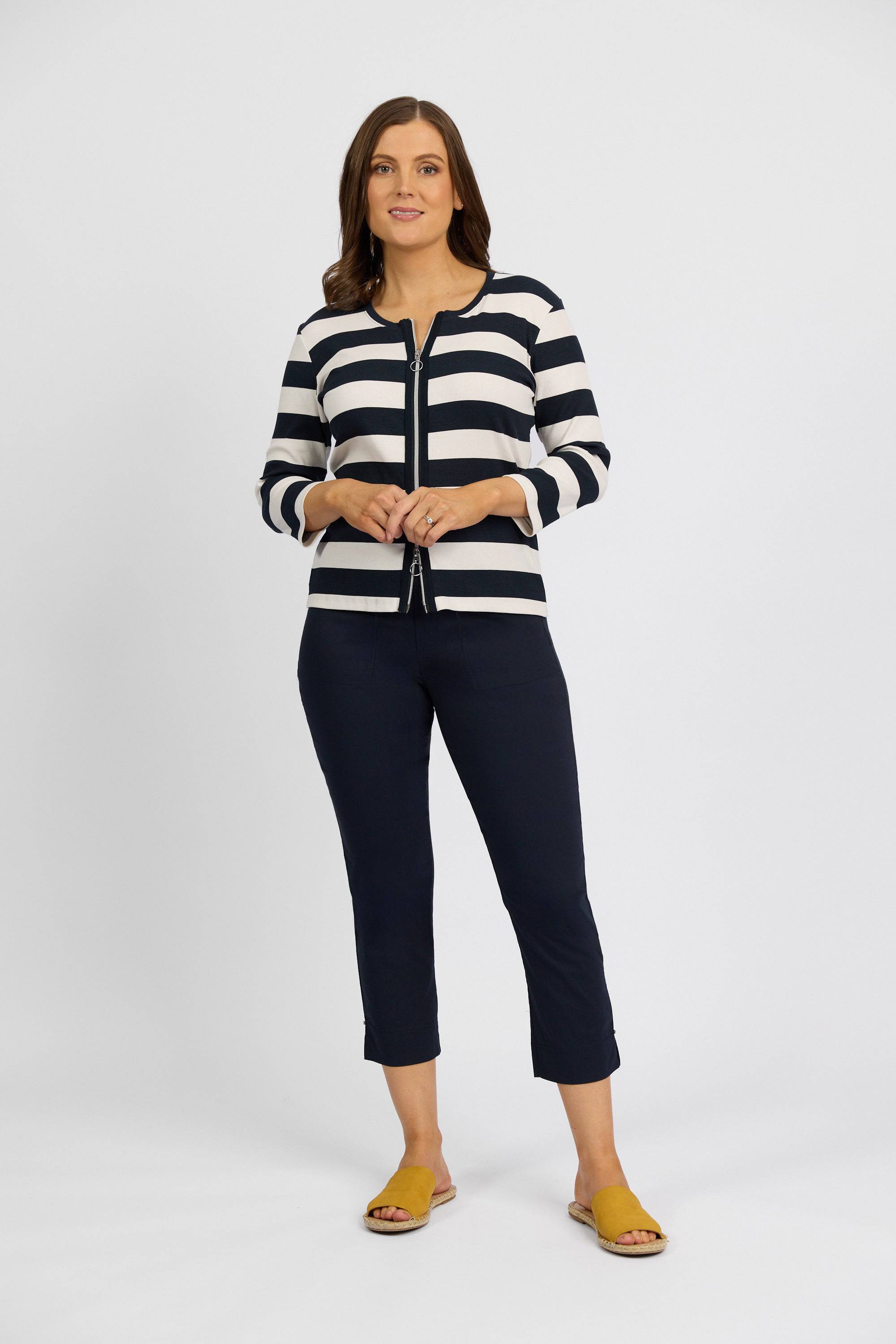 A woman with shoulder-length brown hair and a neutral expression stands against a white background, wearing Vassalli's Paris Tour Capri – Inka pants in dark navy stretch fabric, a black-and-white striped cardigan, and yellow slide sandals.