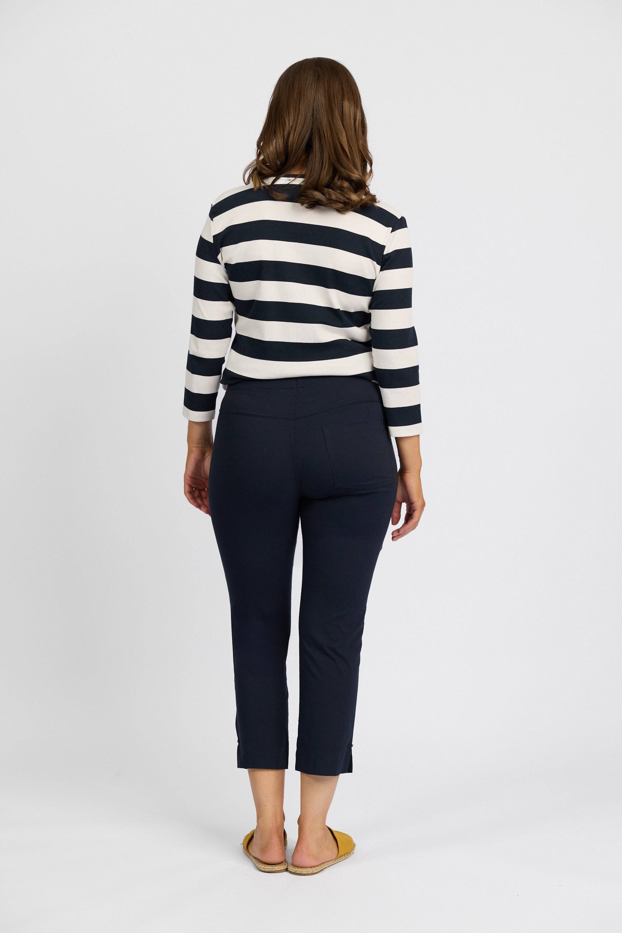 A woman faces away, wearing Vassalli Paris Tour Capri - Inka dark ankle pants, a black and white striped long-sleeve top, and yellow flats, set against a plain white background.