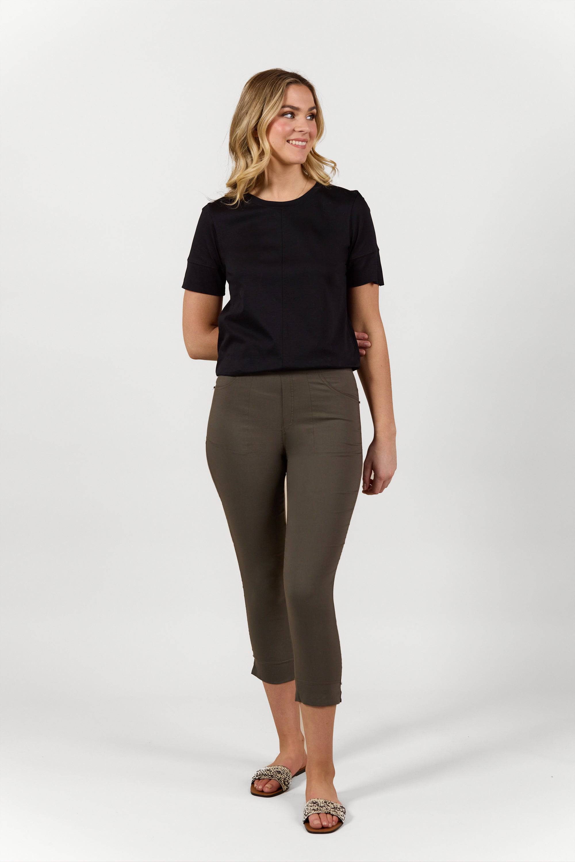A woman with wavy blonde hair wears a short-sleeve black top, Vassalli Paris Tour Capri - Khaki pants, and snakeskin-pattern sandals, standing and smiling against a plain white background.
