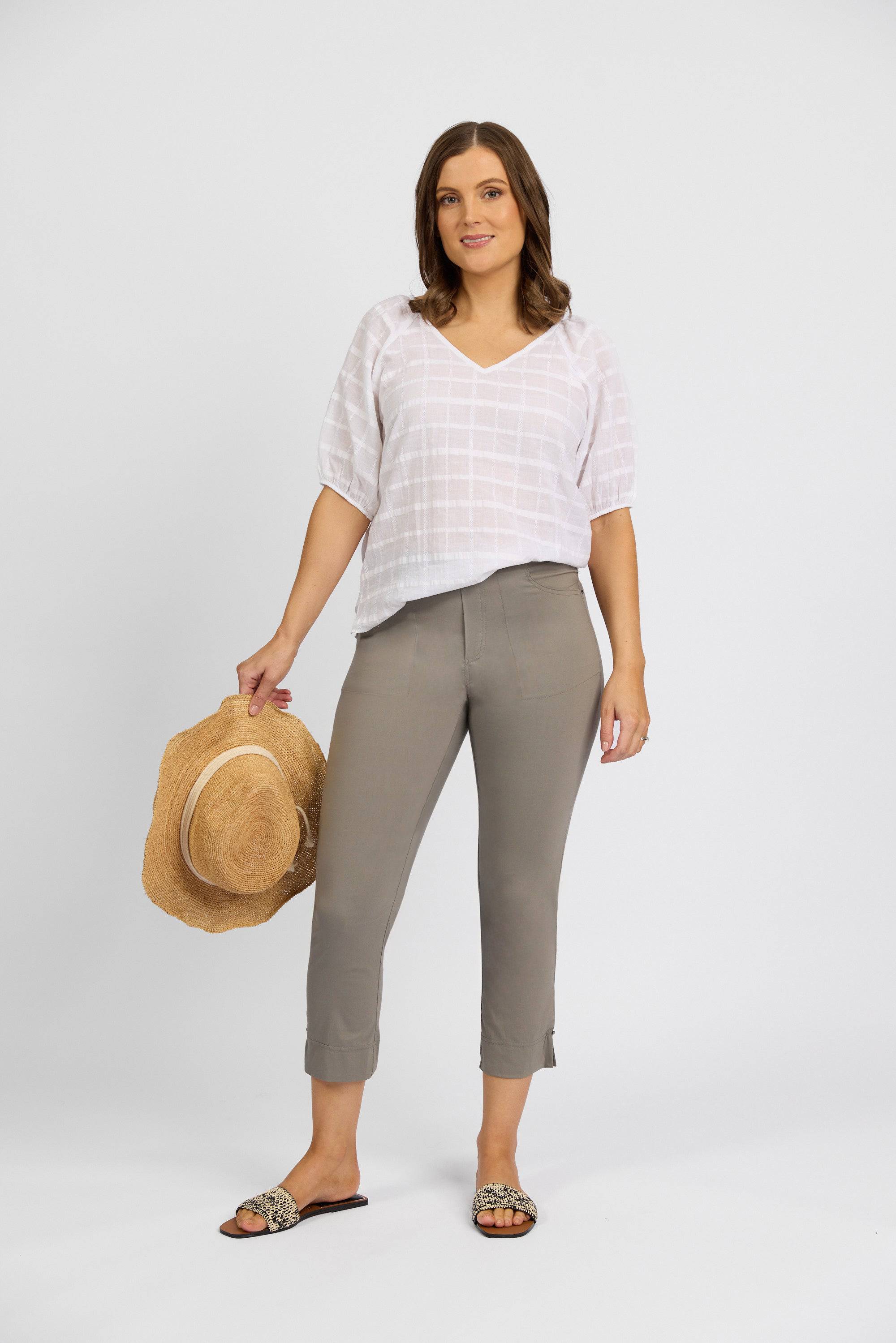 A woman stands against a white background wearing a Vassalli Paris Tour Capri in Latte, a white short-sleeve blouse, and sandals. She holds a straw hat in her left hand and poses in a relaxed, casual manner.