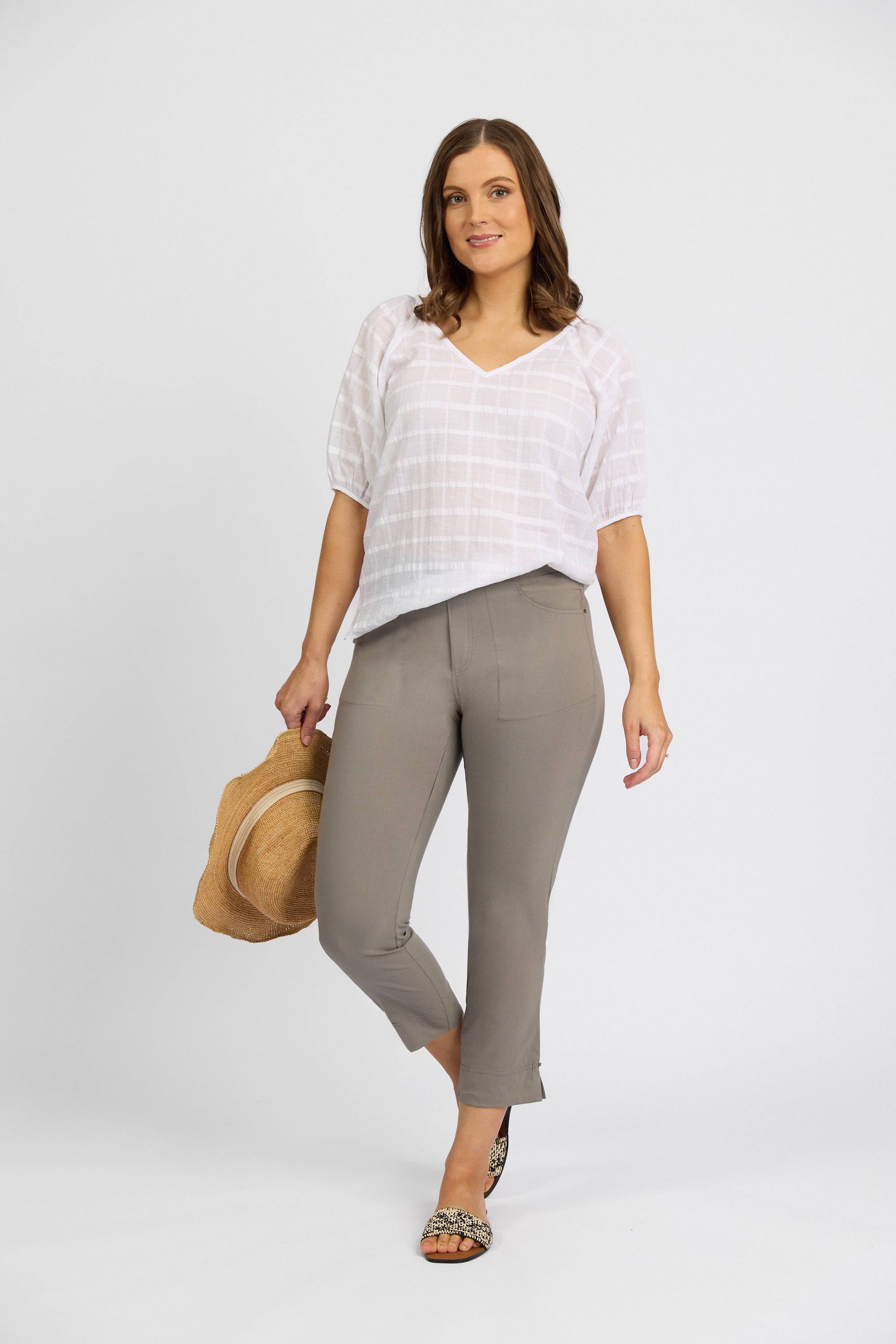 A woman stands against a plain white background, wearing Vassalli’s Paris Tour Capri - Latte, paired with a loose white blouse and sandals. She holds a straw hat in one hand and gently smiles at the camera.