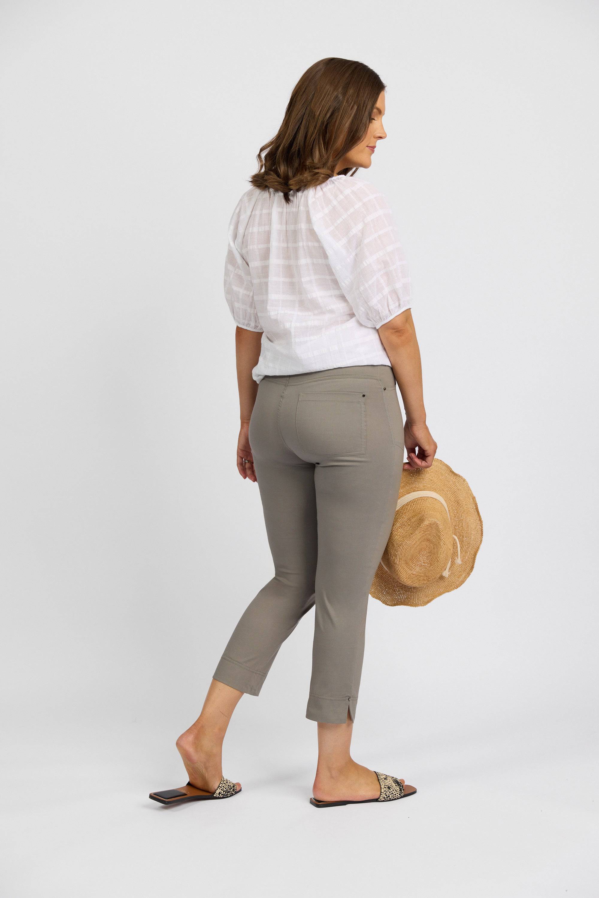 A woman faces away in a white short-sleeve blouse and Vassalli Paris Tour Capri - Latte pants, holding a straw hat. Her shoulder-length brown hair is visible against a plain white background.