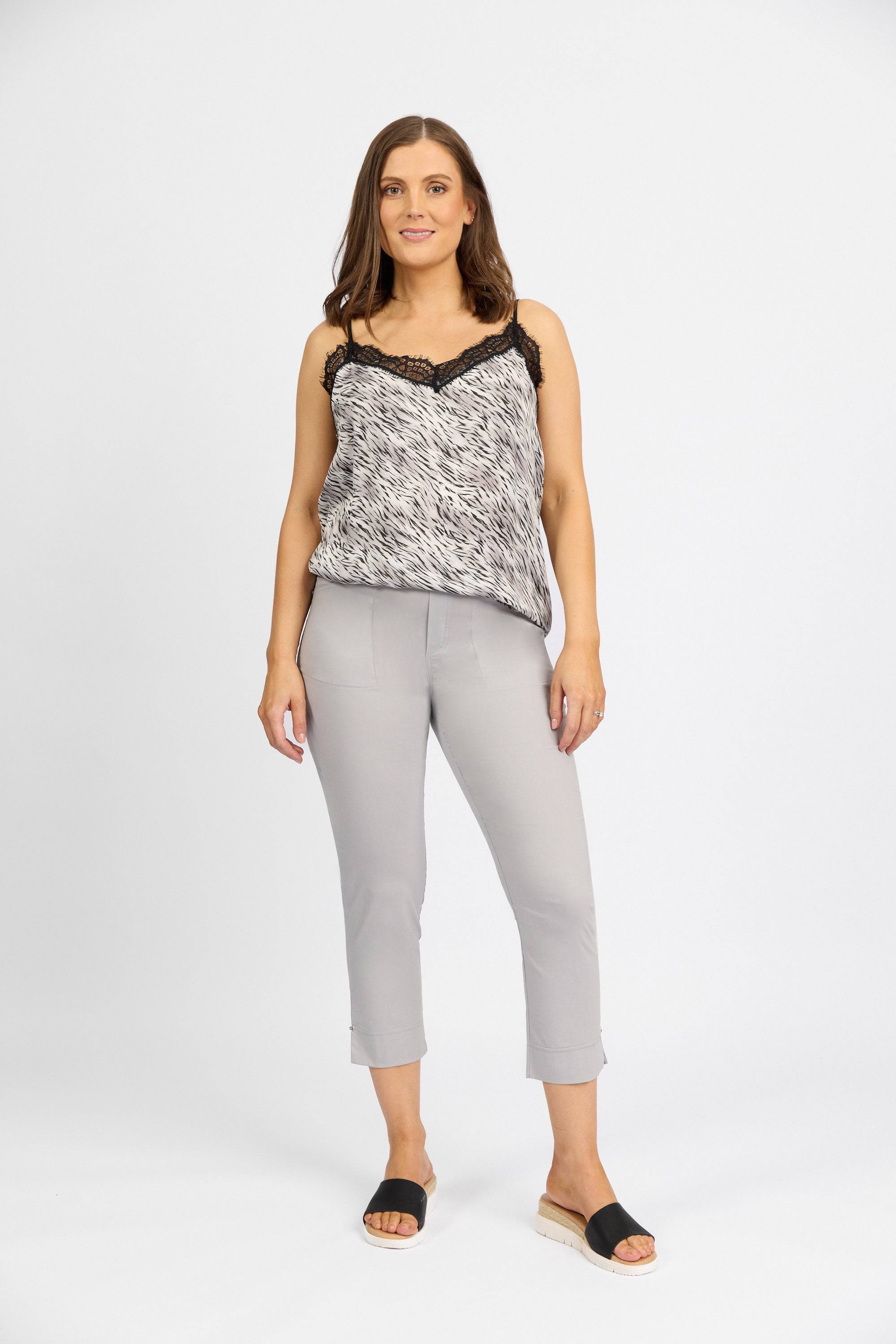 A woman with shoulder-length brown hair and a neutral expression wears a sleeveless patterned top with lace trim, Vassalli Paris Tour Capri pants in Silver, and black slide sandals against a plain white background.