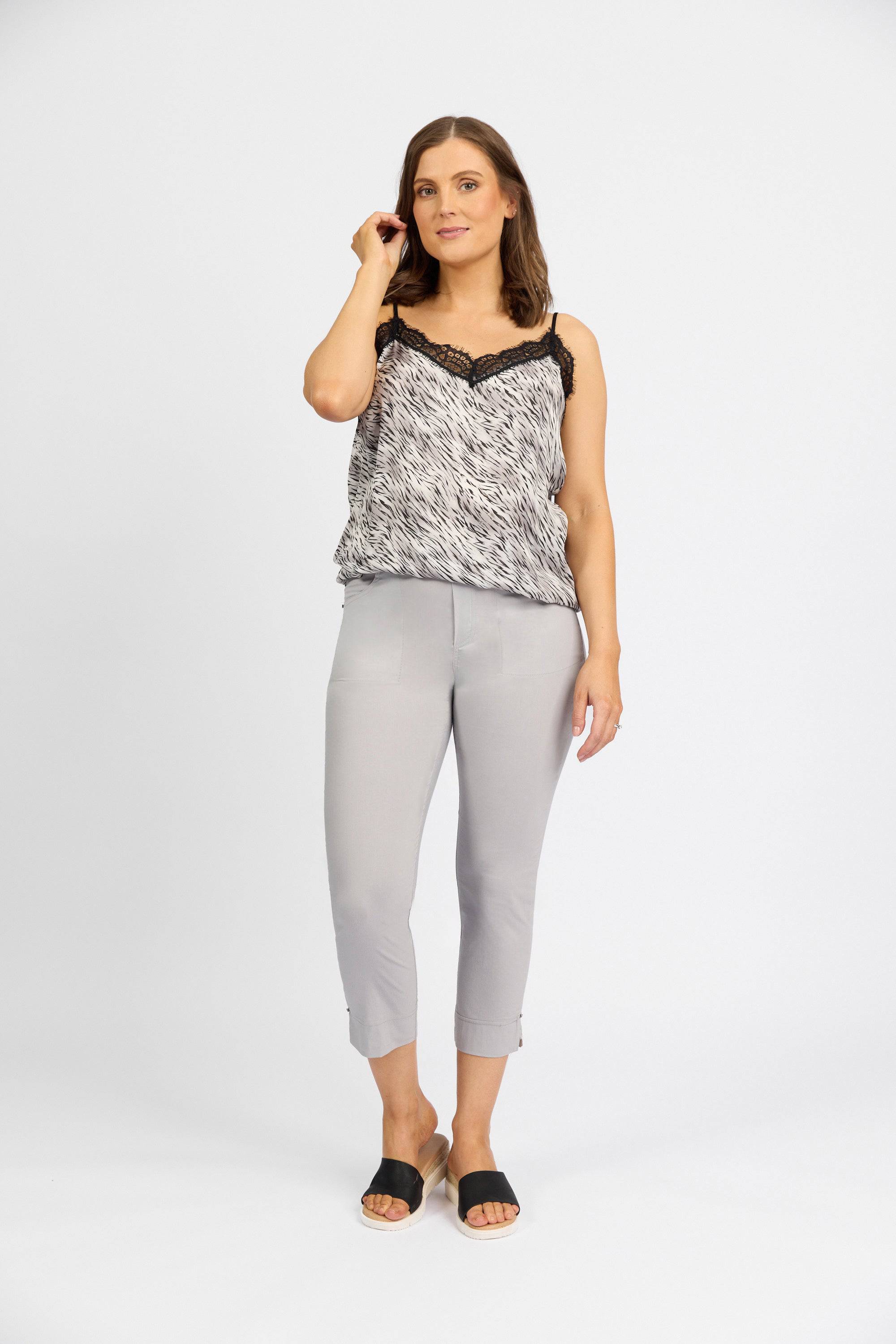 A woman with shoulder-length brown hair and a neutral expression stands against a white background, wearing a patterned sleeveless lace-trim top, Vassalli Paris Tour Capri - Silver cropped pants in stretch fabric, and black slide sandals.
