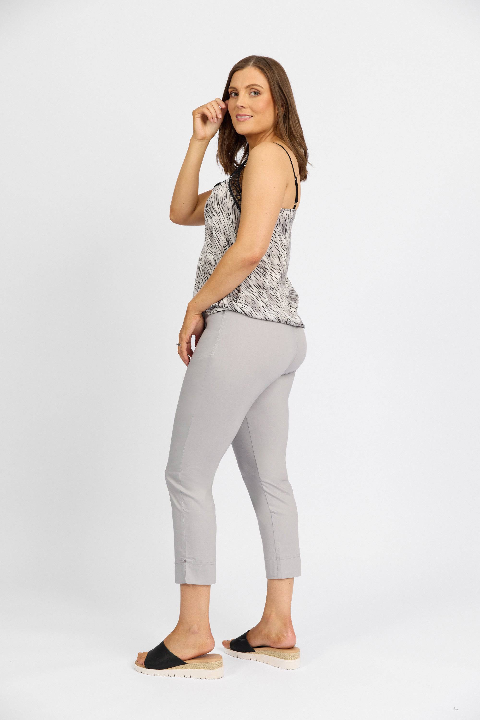 A woman stands sideways against a white background, wearing a patterned sleeveless top and Vassalli Paris Tour Capri - Silver pants, crafted from stretch fabric, with black slide sandals. She looks at the camera, one hand touching her hair.