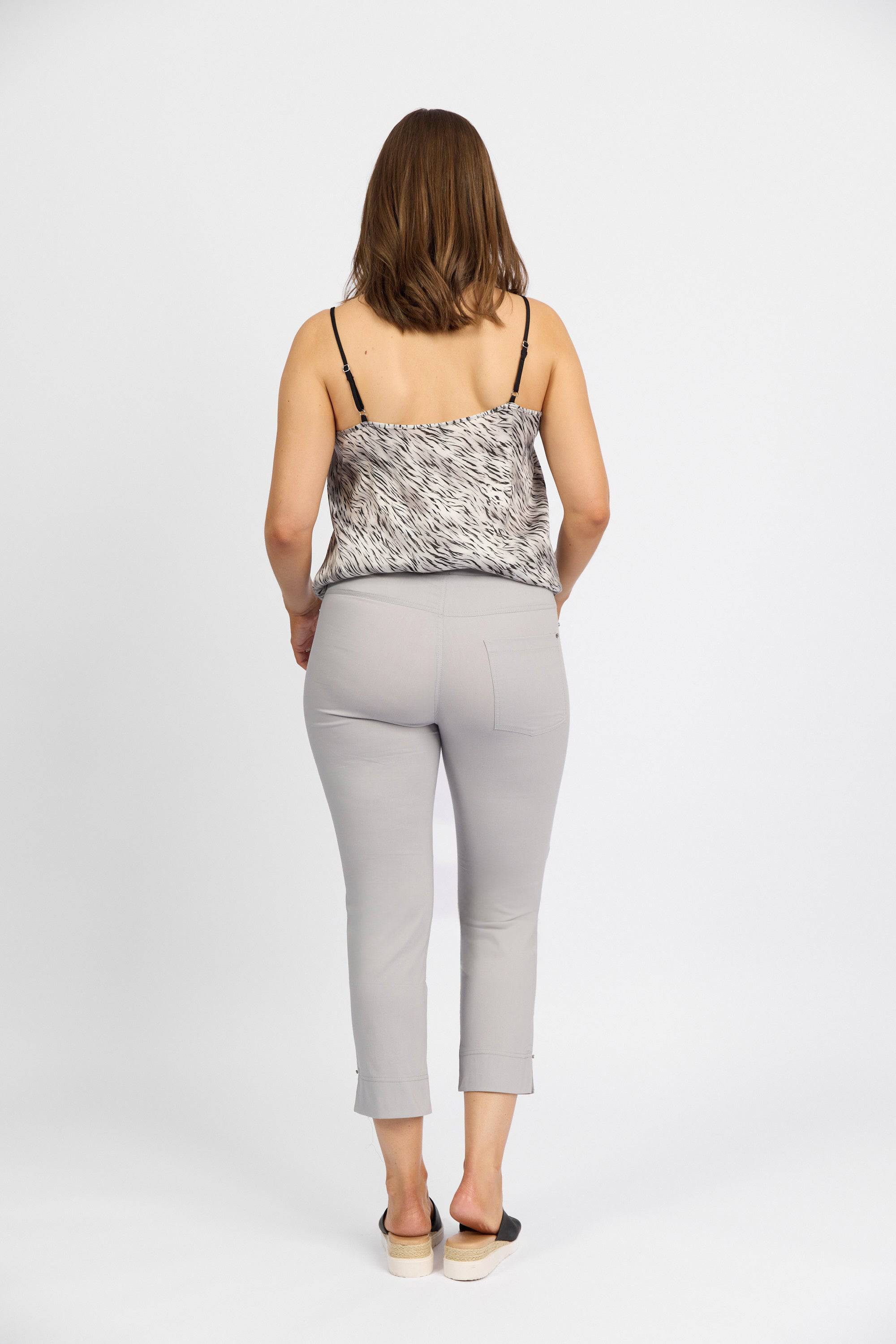 A woman faces away from the camera, dressed in a black-and-white sleeveless top, Vassalli Paris Tour Capri pants in Silver made from stretch fabric, and black slip-on shoes, all set against a plain white background.