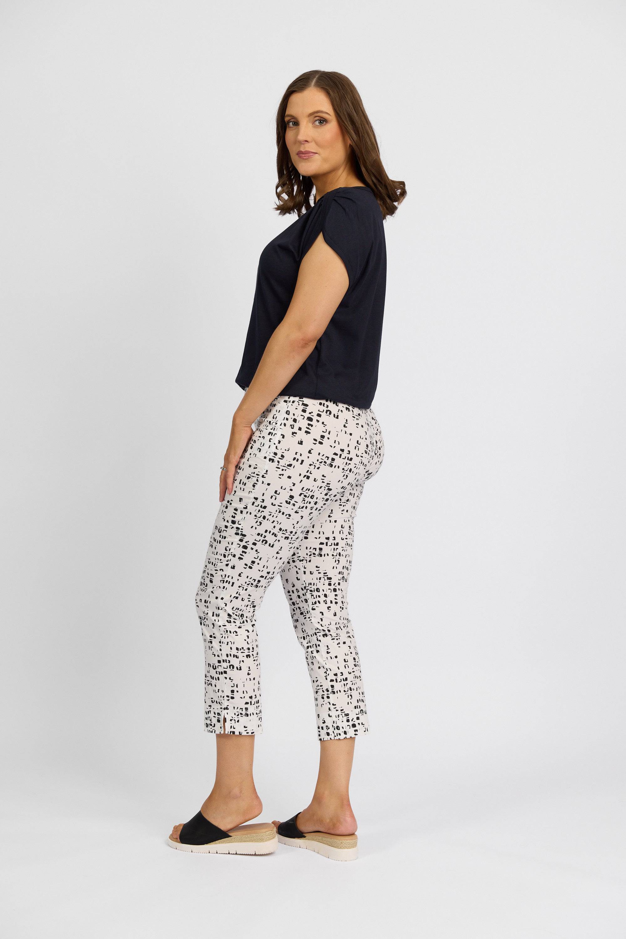 A woman stands sideways against a plain background, wearing a navy short-sleeve top, Vassalli Paris Printed Tour Capri - Tonic pants in white with a black pattern, and black-and-beige slide sandals.