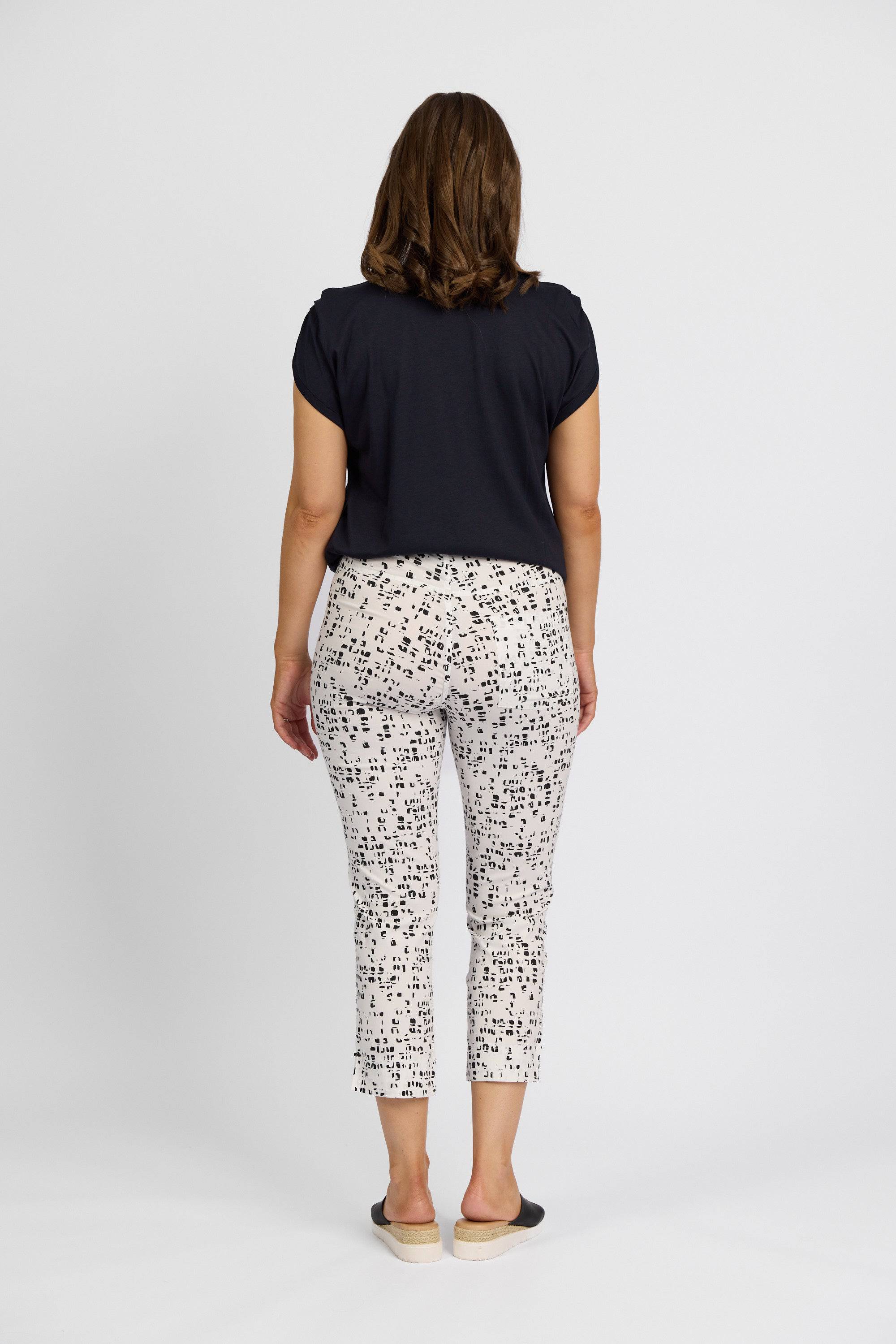 A woman faces away from the camera, wearing a Vassalli short-sleeve black shirt, Vassalli Paris Printed Tour Capri - Tonic in stretch fabric with a black dot pattern, and black slip-on shoes against a plain white background.