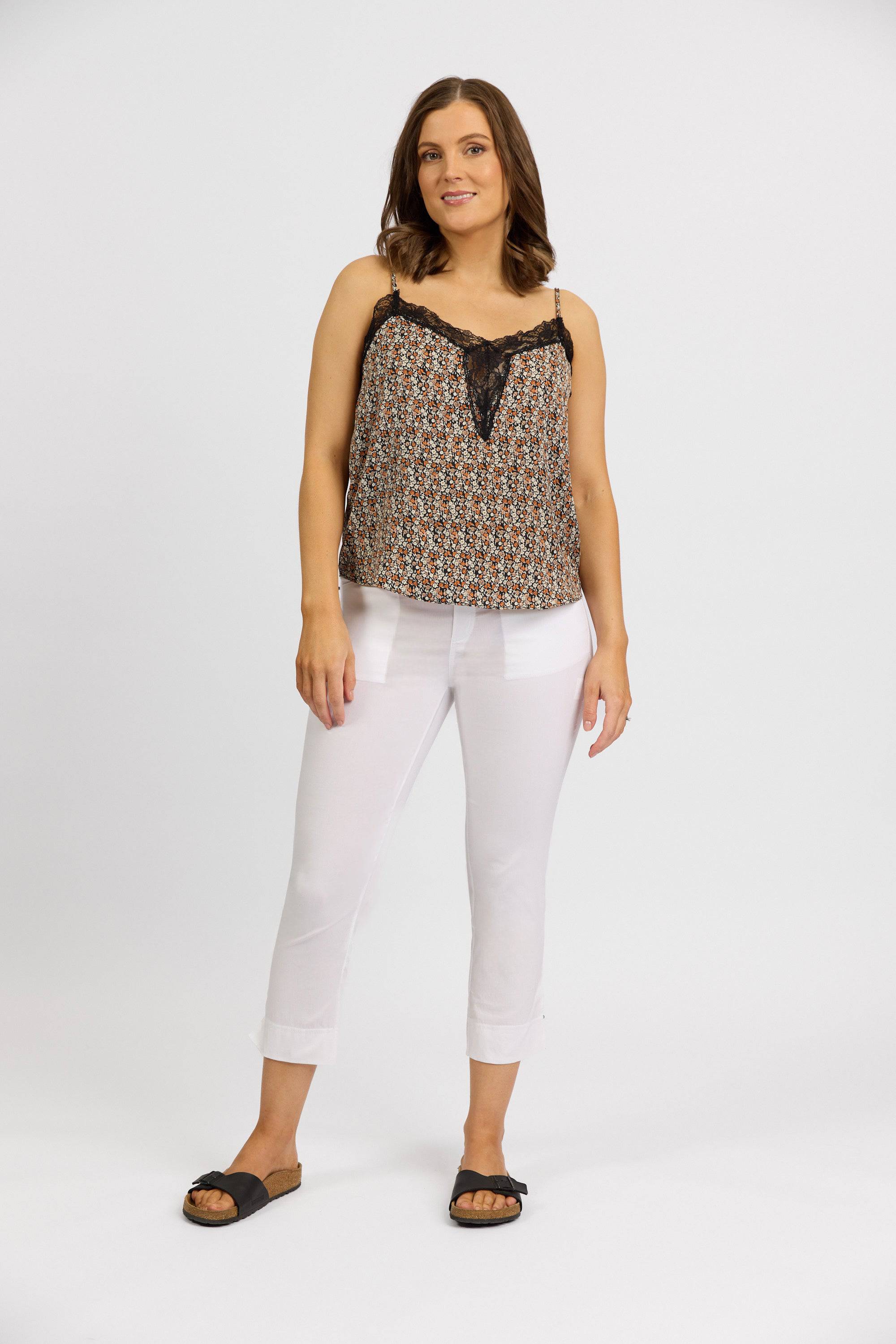 A woman with shoulder-length brown hair smiles softly, wearing a Vassalli patterned sleeveless lace-trim top, Vassalli Paris Tour Capri - White pants, and black sandals against a plain white background.