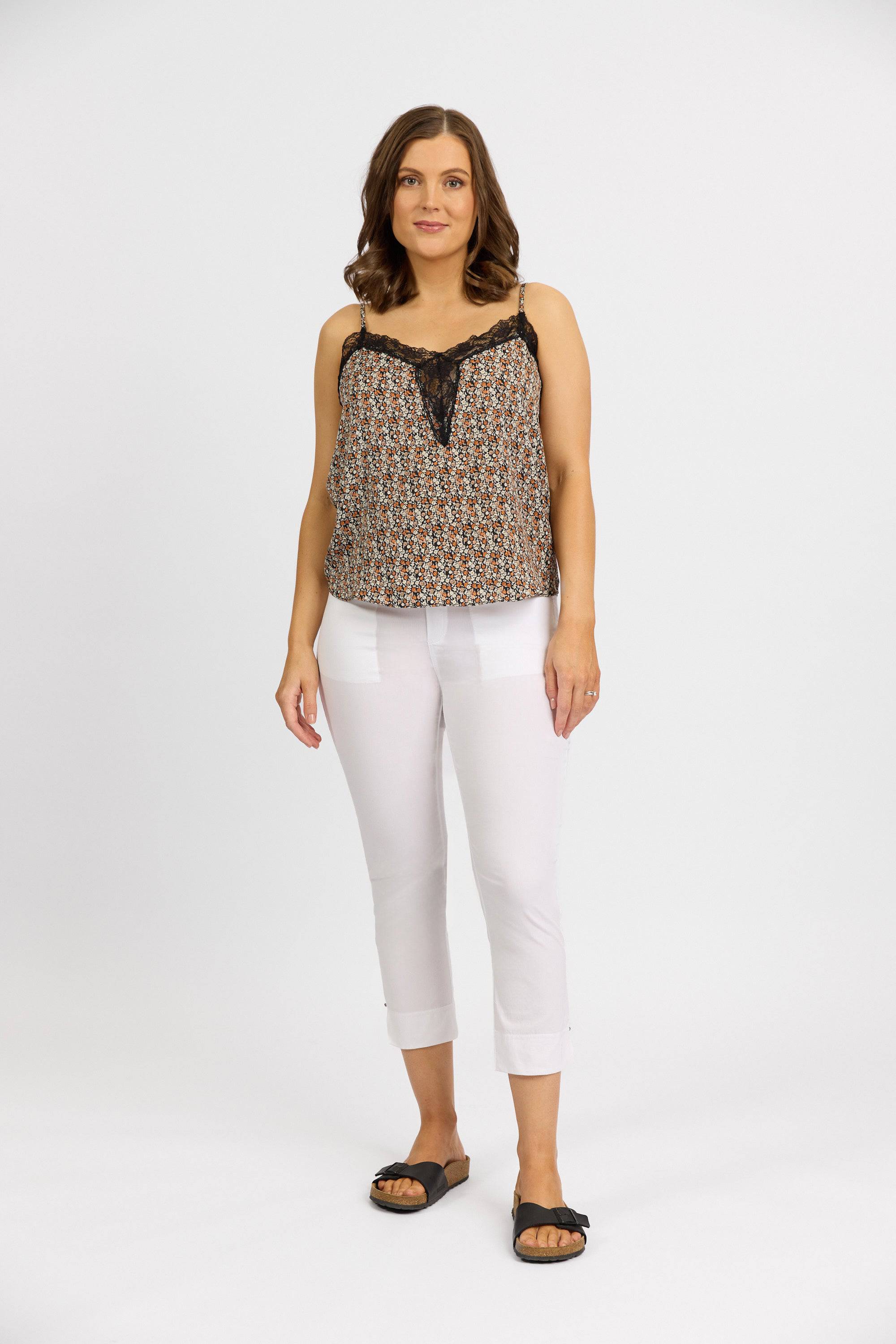 A woman with shoulder-length brown hair stands against a white background, wearing a sleeveless patterned top, Vassalli Paris Tour Capri - White pants, and black sandals. She looks at the camera with a neutral expression.