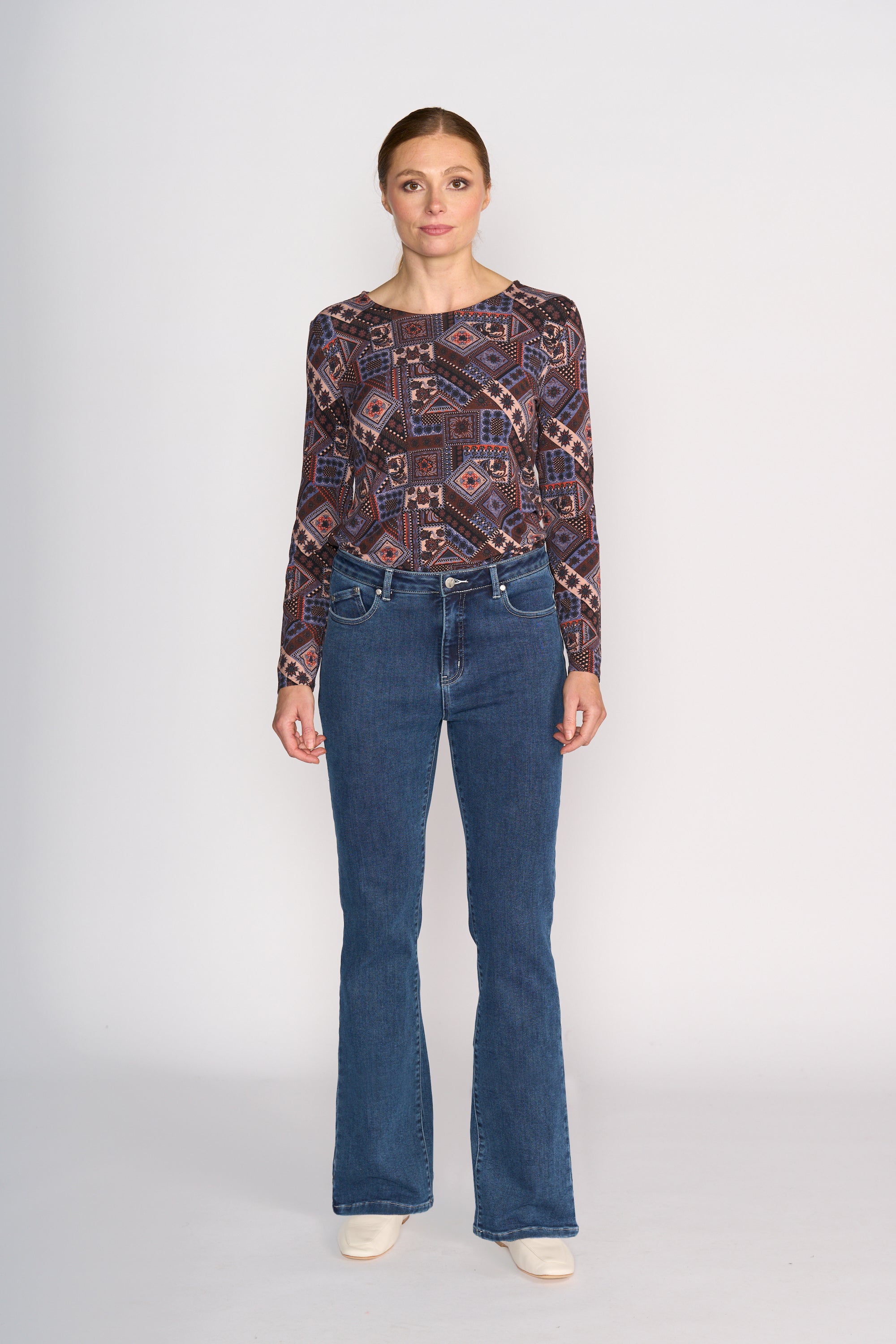 A woman stands facing forward against a plain background, wearing a patterned long-sleeve top, white shoes, and Vassalli's Theory Jean - Dark Blue Brushed.