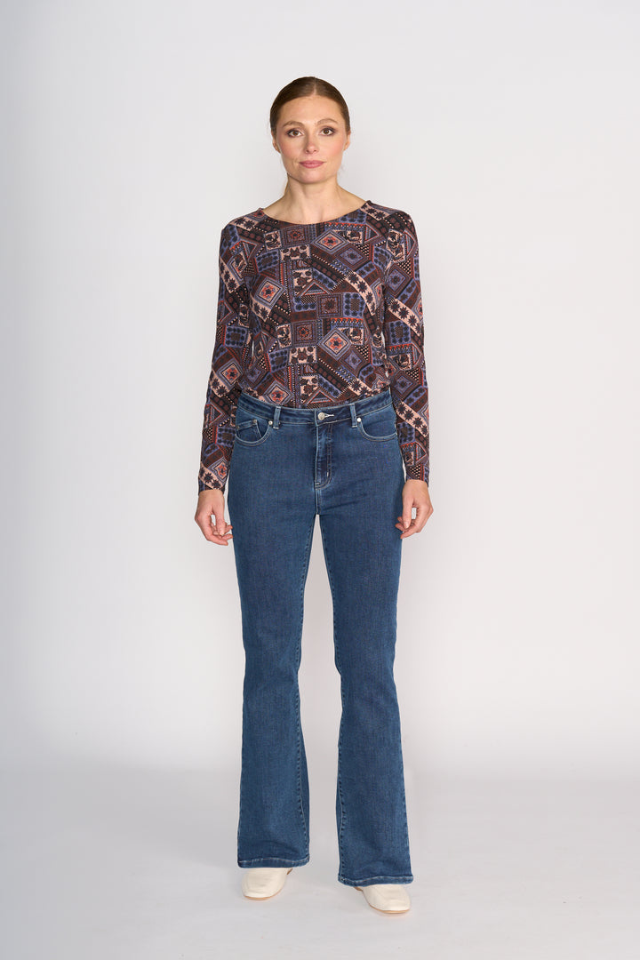 A woman stands facing forward against a plain background, wearing a patterned long-sleeve top, white shoes, and Vassalli's Theory Jean - Dark Blue Brushed.