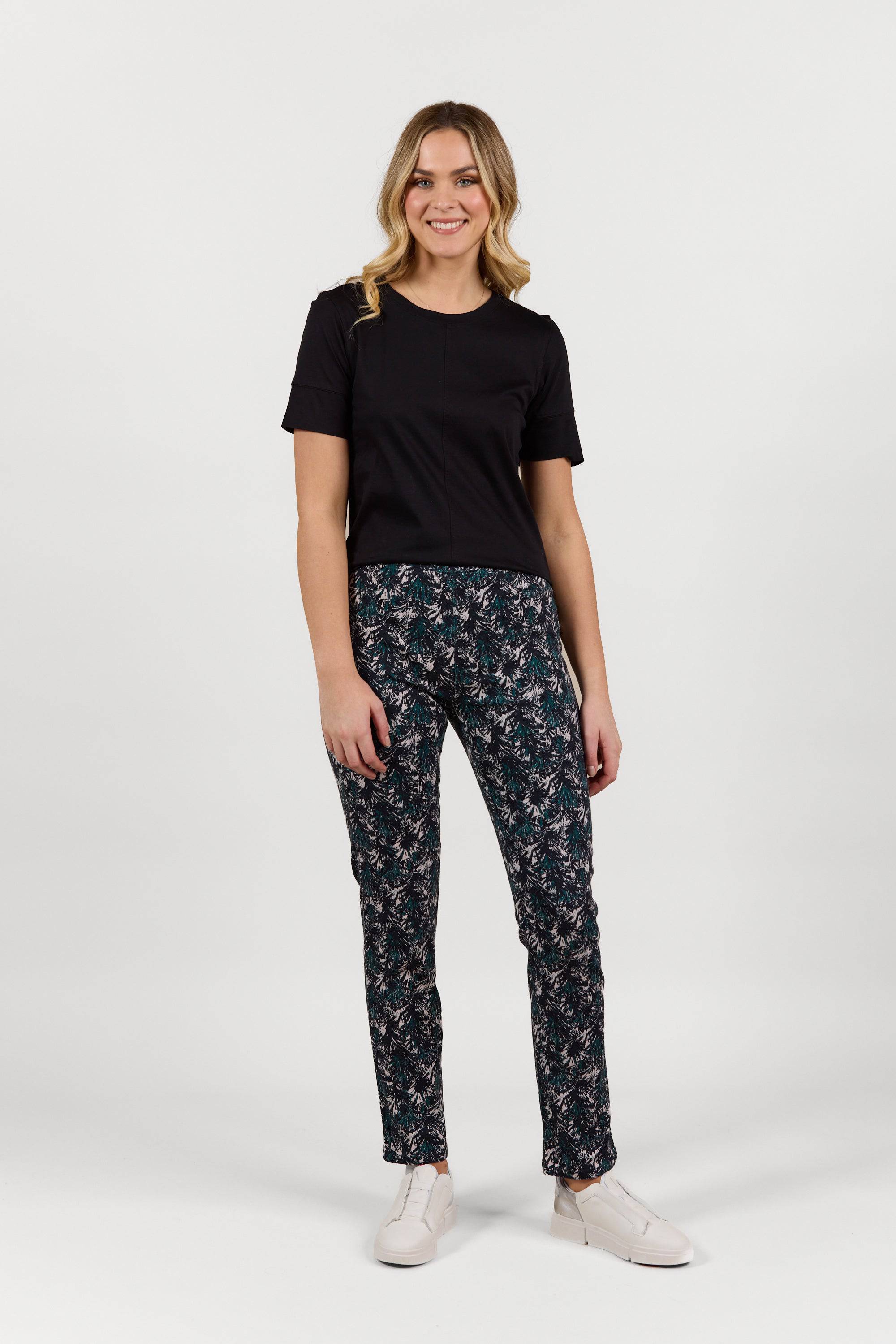 A woman with wavy blonde hair smiles while wearing a black short-sleeve top, Vassalli Bay Pull On - Aspen full-length printed pants, and white sneakers, standing against a plain white background.