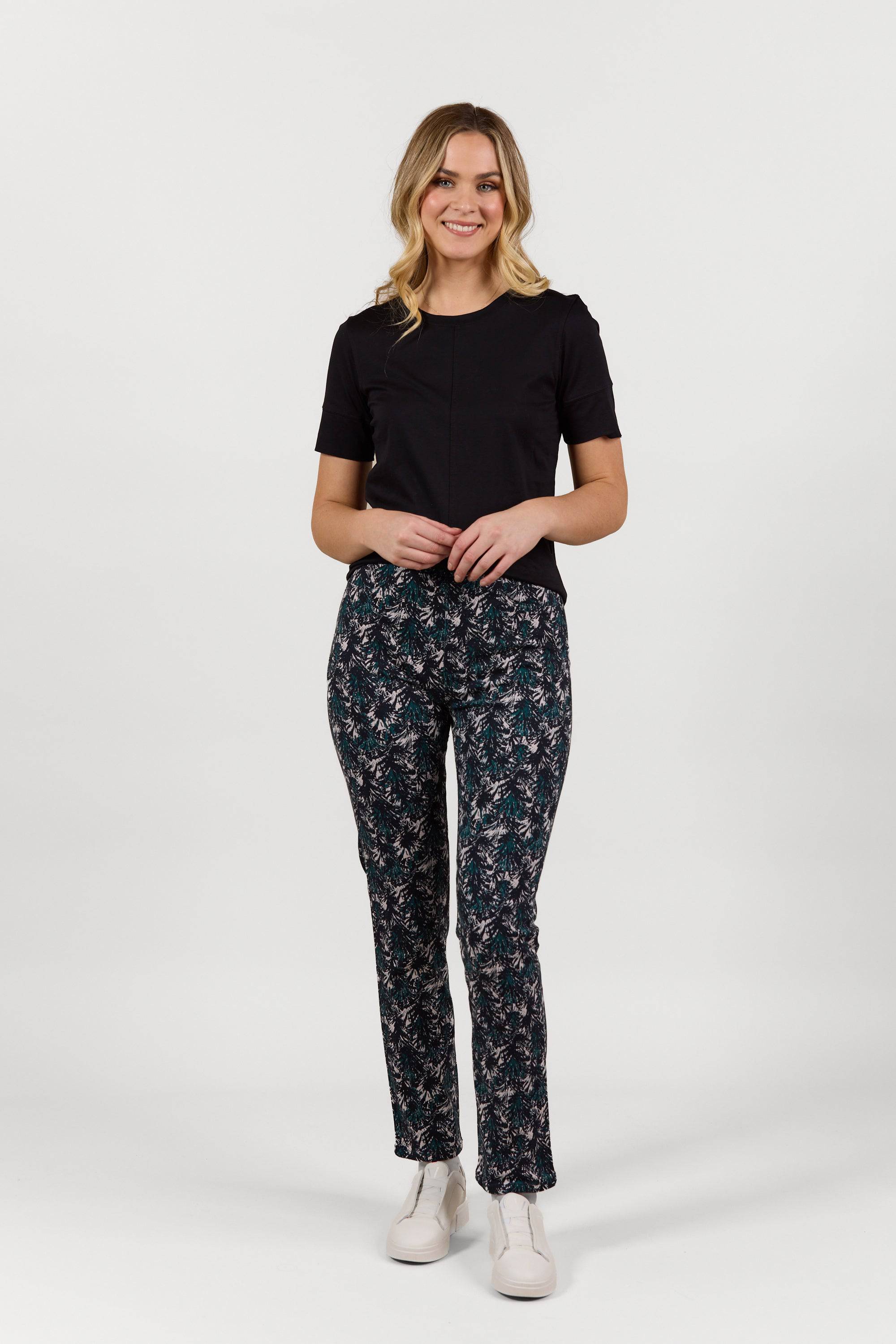 A woman with light wavy hair stands smiling against a plain white background, wearing a black short-sleeve top, Vassalli Bay Pull On - Aspen full length printed pants, and white sneakers, with her hands clasped in front.