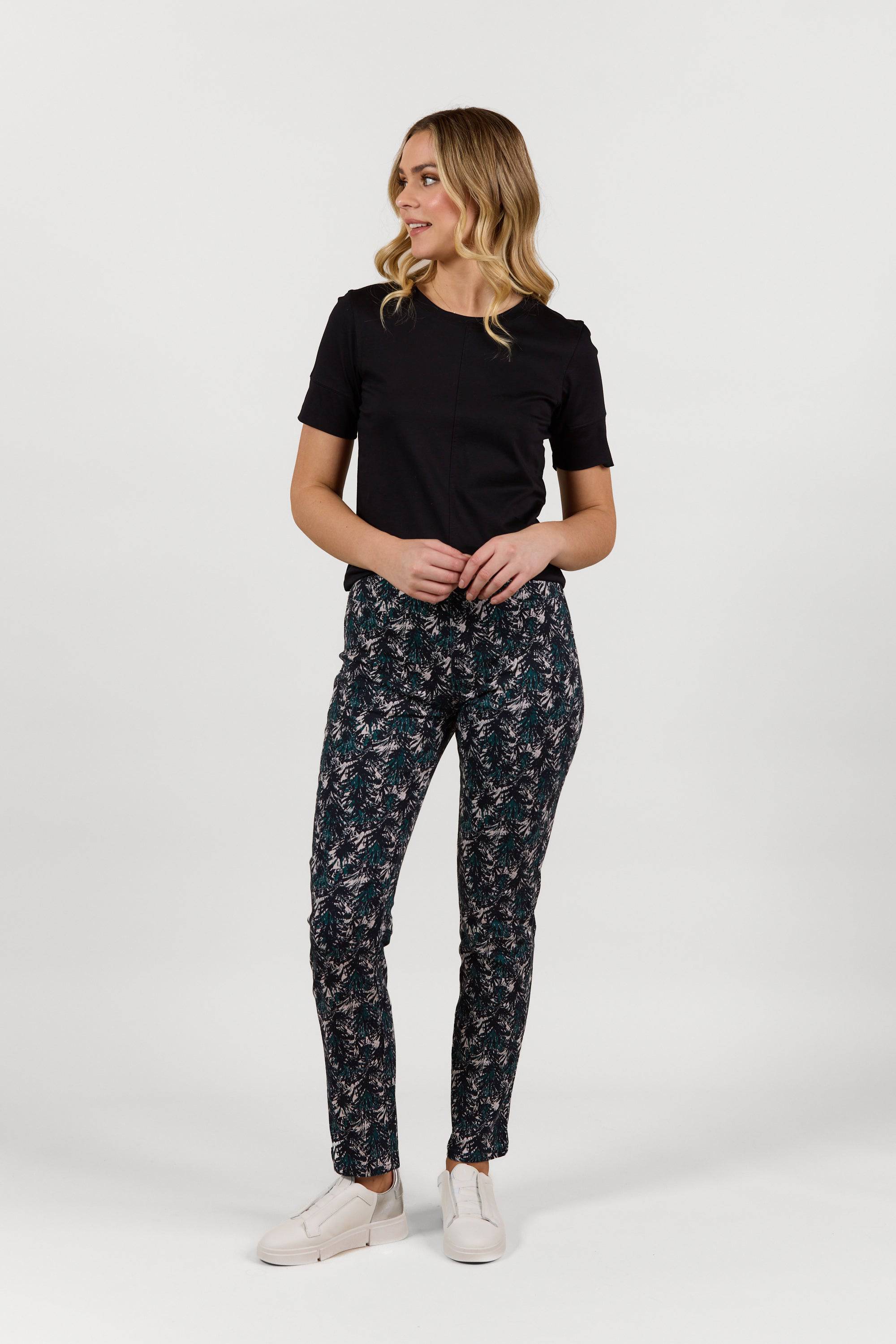 A woman with wavy, shoulder-length blonde hair wears the Vassalli Bay Pull On - Aspen full-length floral pants, a black short-sleeve top, and white shoes against a plain white background, looking to her left.