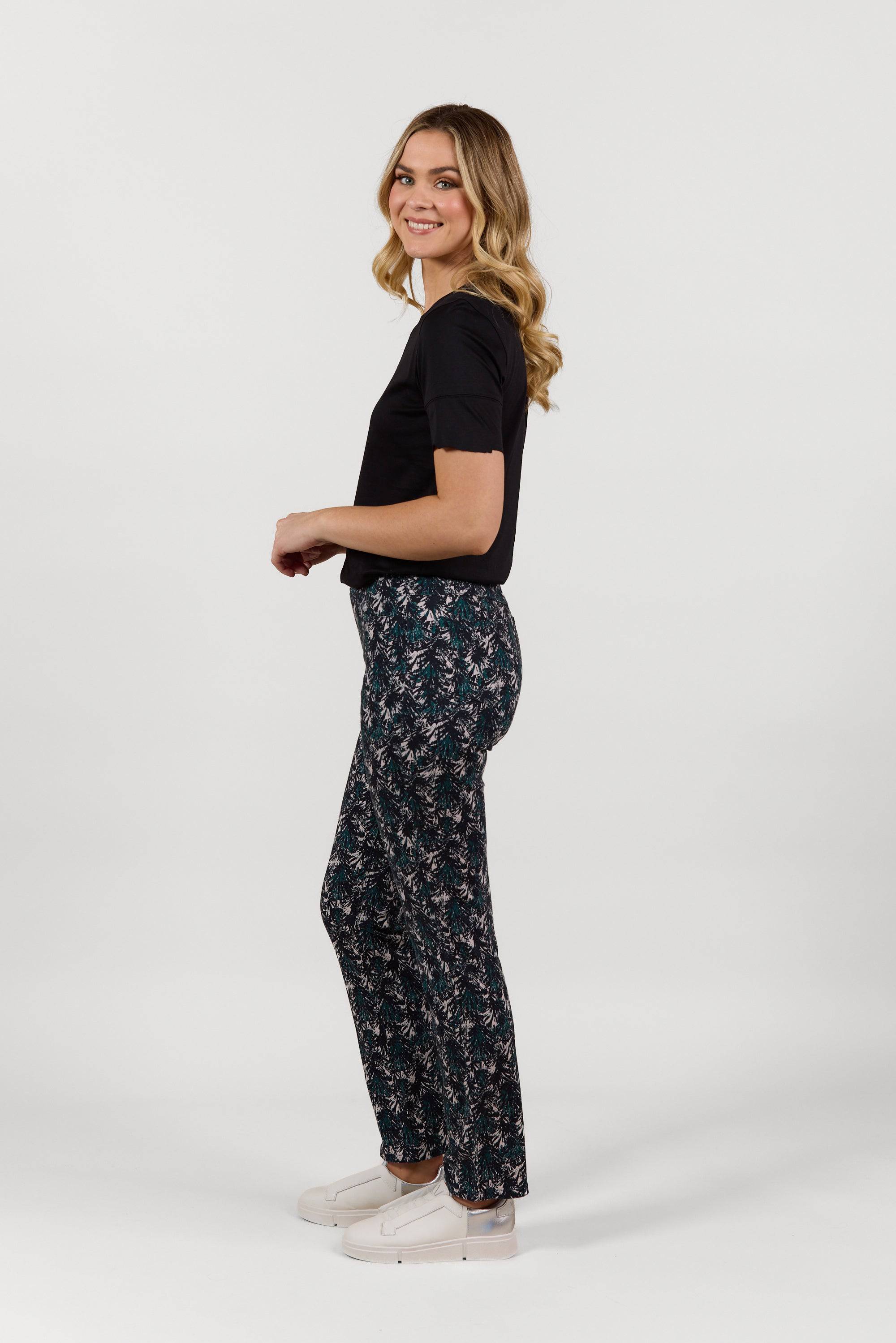 A woman with wavy blonde hair stands sideways, smiling. She is wearing a black t-shirt, Vassalli Bay Pull On - Aspen full length printed pants with a floral design, and white slip-on shoes against a plain white background.