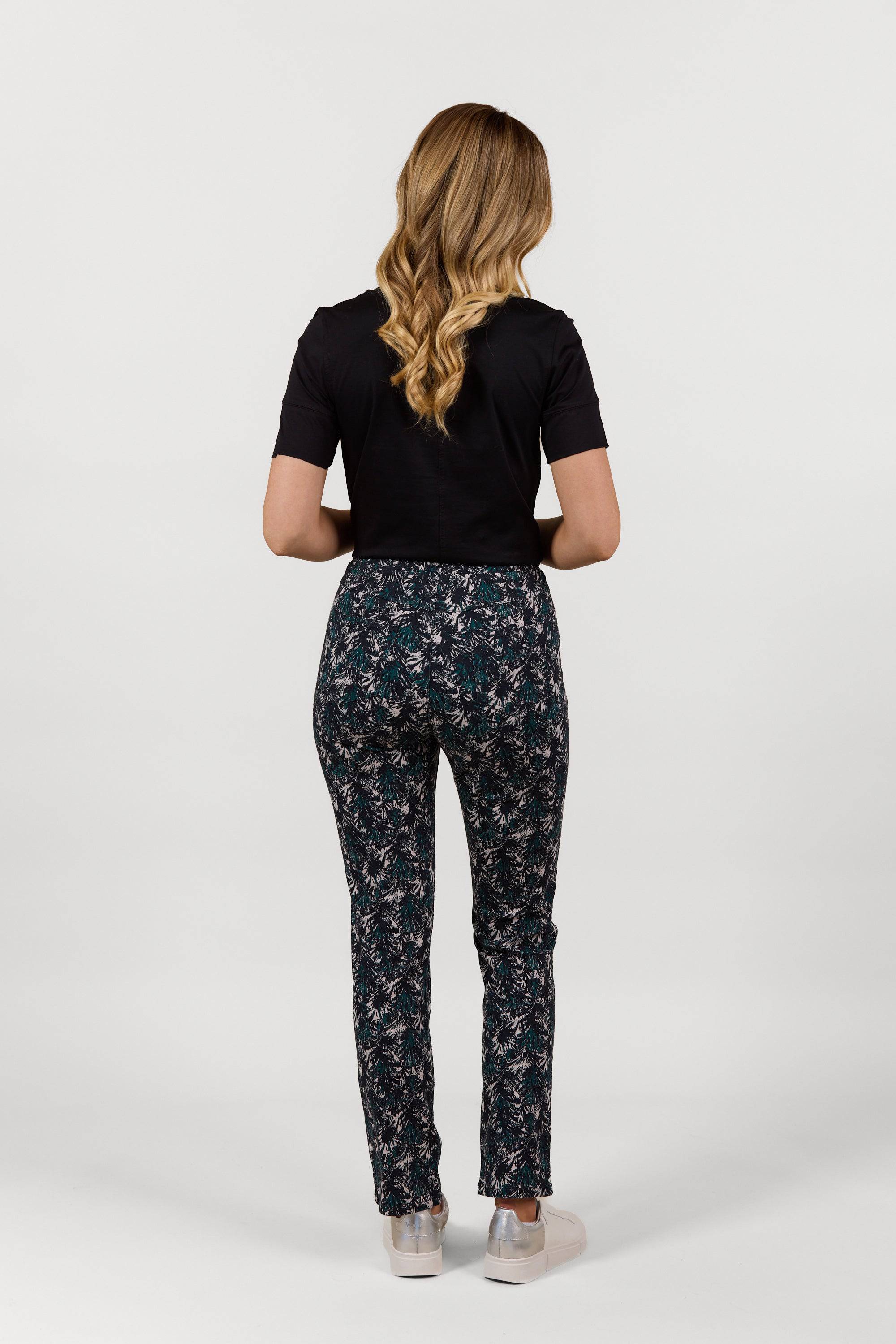 A person with long wavy hair, wearing a black short-sleeve top, Vassalli Bay Pull On - Aspen full-length printed pants, and white shoes stands facing away against a plain white background.