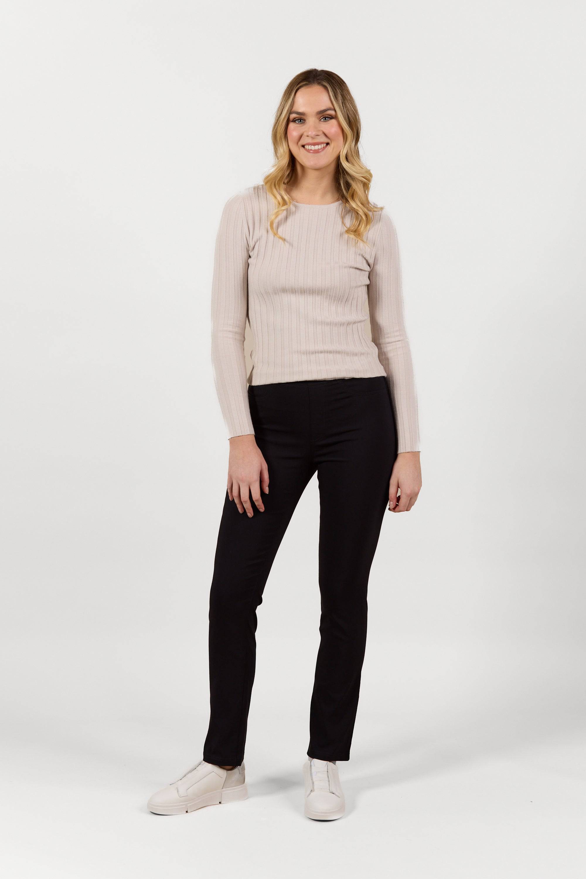 A woman with long blonde hair, in a light long-sleeve top, white shoes, and Vassalli Bay Pull On - Black Bliss pants, stands smiling against a plain white background.