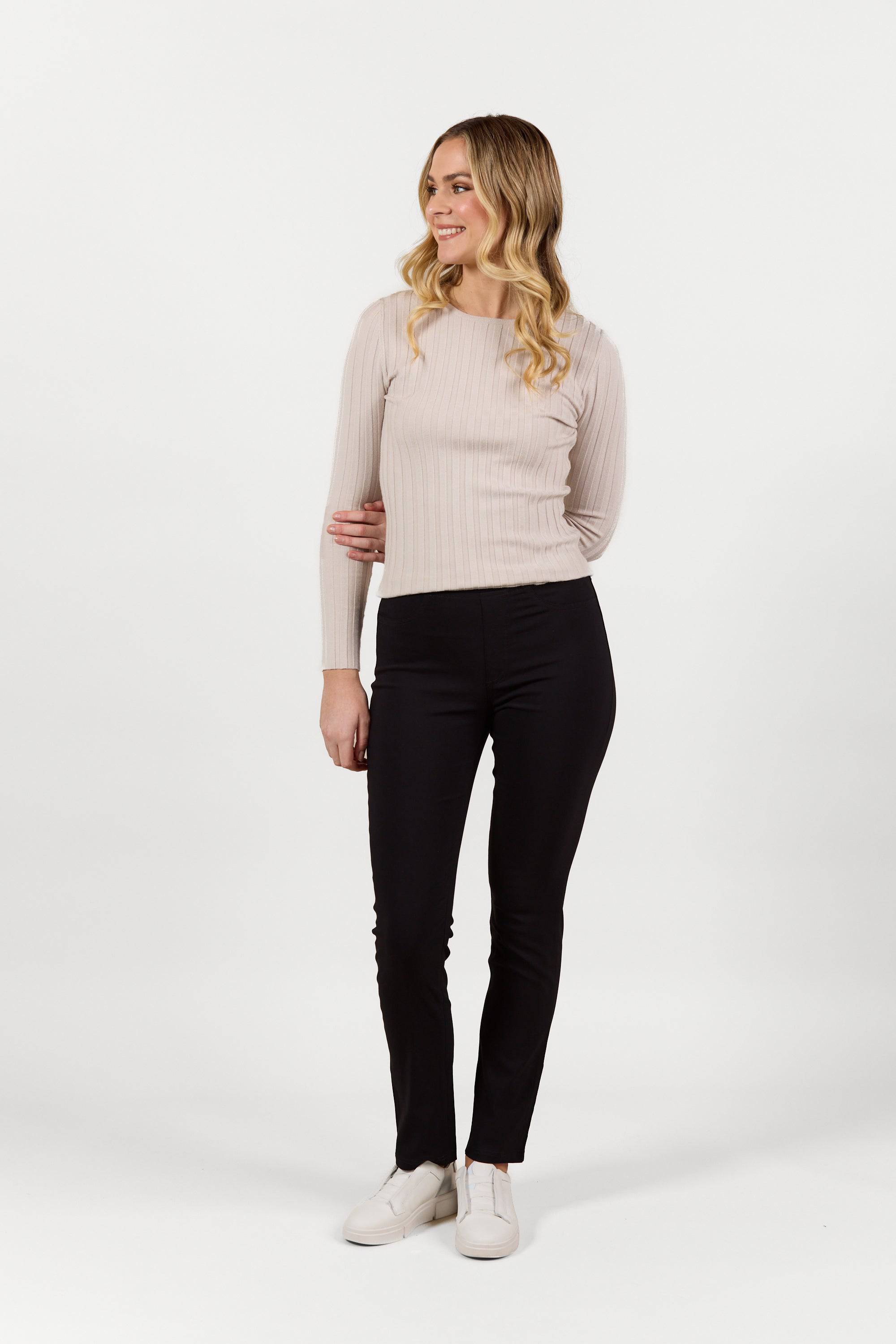 A smiling woman with long blonde wavy hair models Vassalli’s Bay Pull On - Black Bliss pants, paired with a light beige ribbed long-sleeve top and white shoes, standing against a plain white background and looking to her left.