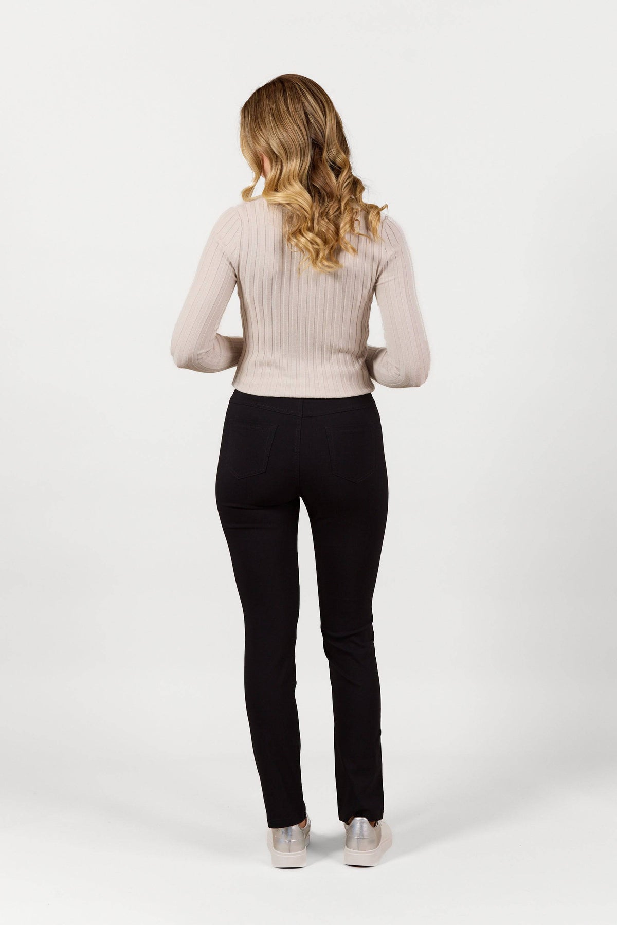 A woman with long wavy blonde hair stands facing away, wearing a light beige ribbed sweater, Vassalli Bay Pull On - Black Bliss slim leg pants, and silver shoes against a plain white background.