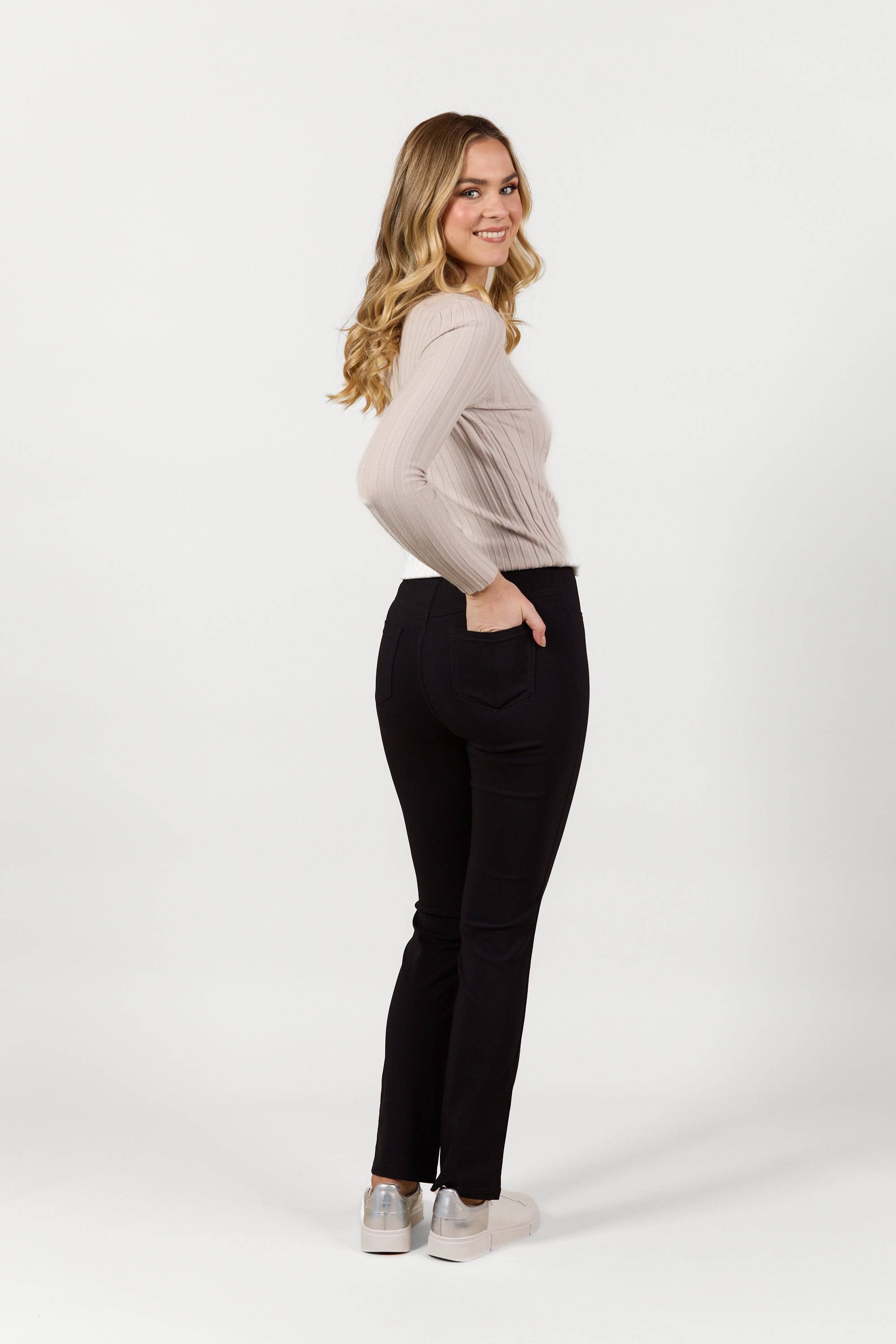 A woman with long, wavy blonde hair smiles confidently in Vassalli's Bay Pull On - Black Bliss pants, pairing them with a beige long-sleeve top and white sneakers, posing sideways against a plain white background.