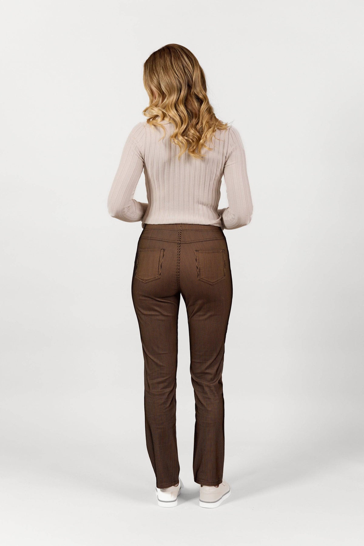 A woman with wavy blonde hair faces away, wearing a light beige ribbed sweater, Vassalli Bay Pull On - Bronze Velvet pants, and white sneakers against a plain white background.