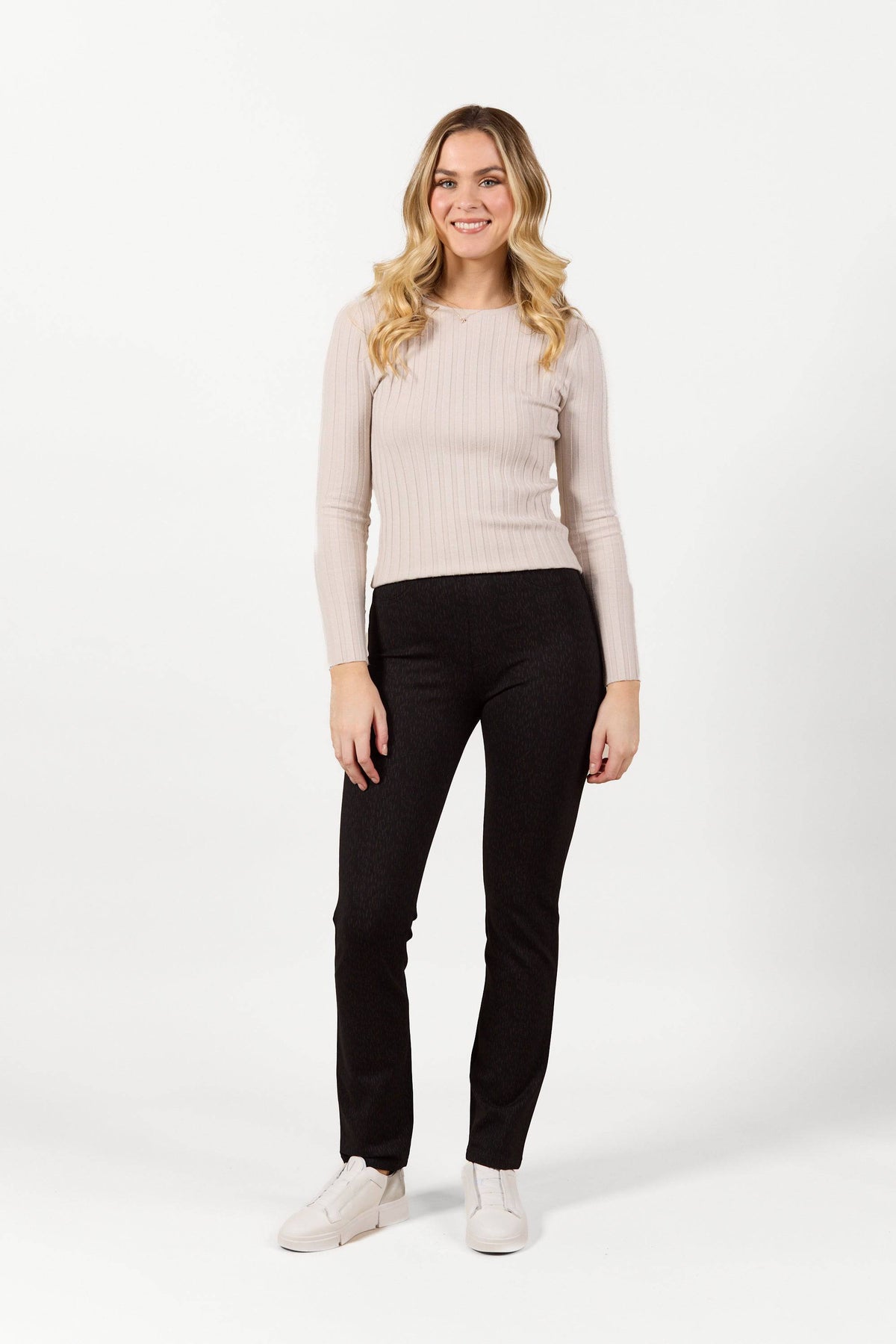 A woman with long, wavy blonde hair smiles against a plain white background, wearing the Vassalli Bay Pull On - Comet slim leg pants in black, a light beige ribbed long-sleeve top, and white shoes.