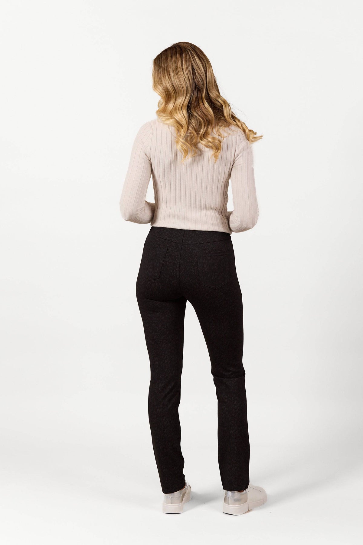A woman with wavy blonde hair stands with her back to the camera, wearing Vassalli's Bay Pull On - Comet dark slim leg pants, a light beige ribbed sweater, and white shoes against a plain white background.