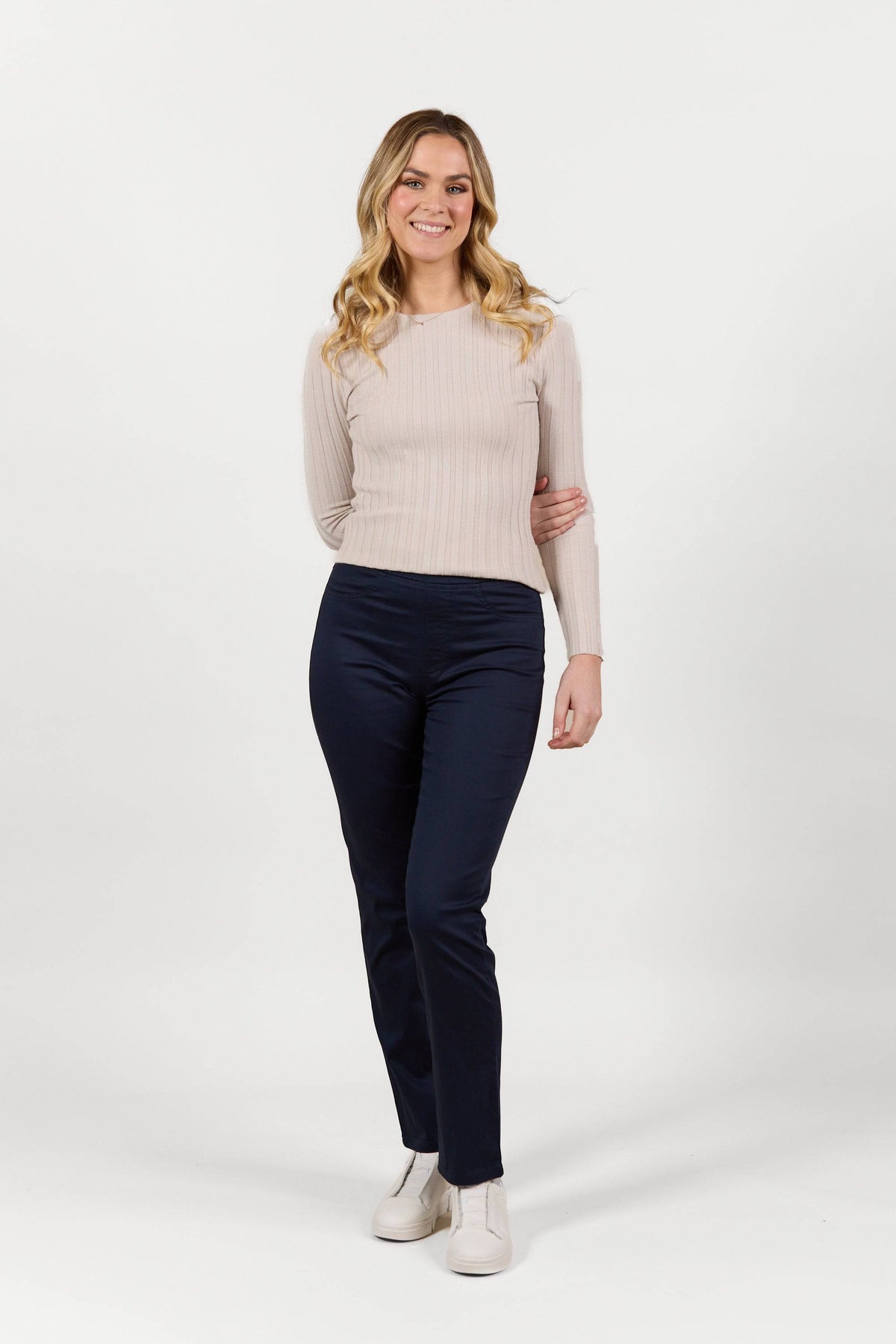 A woman with long blonde hair, wearing a light beige ribbed sweater, Vassalli Bay Pull On - Navy pants, and white shoes stands smiling in front of a plain white background.