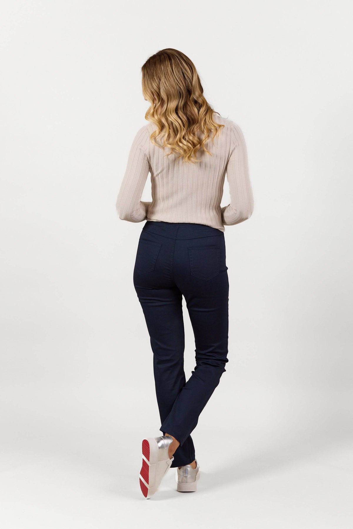 A woman with wavy blonde hair stands facing away, wearing a light beige ribbed sweater and Vassalli Bay Pull On - Navy slim leg pants made from stretch fabric, paired with white shoes with red soles, against a plain white background.