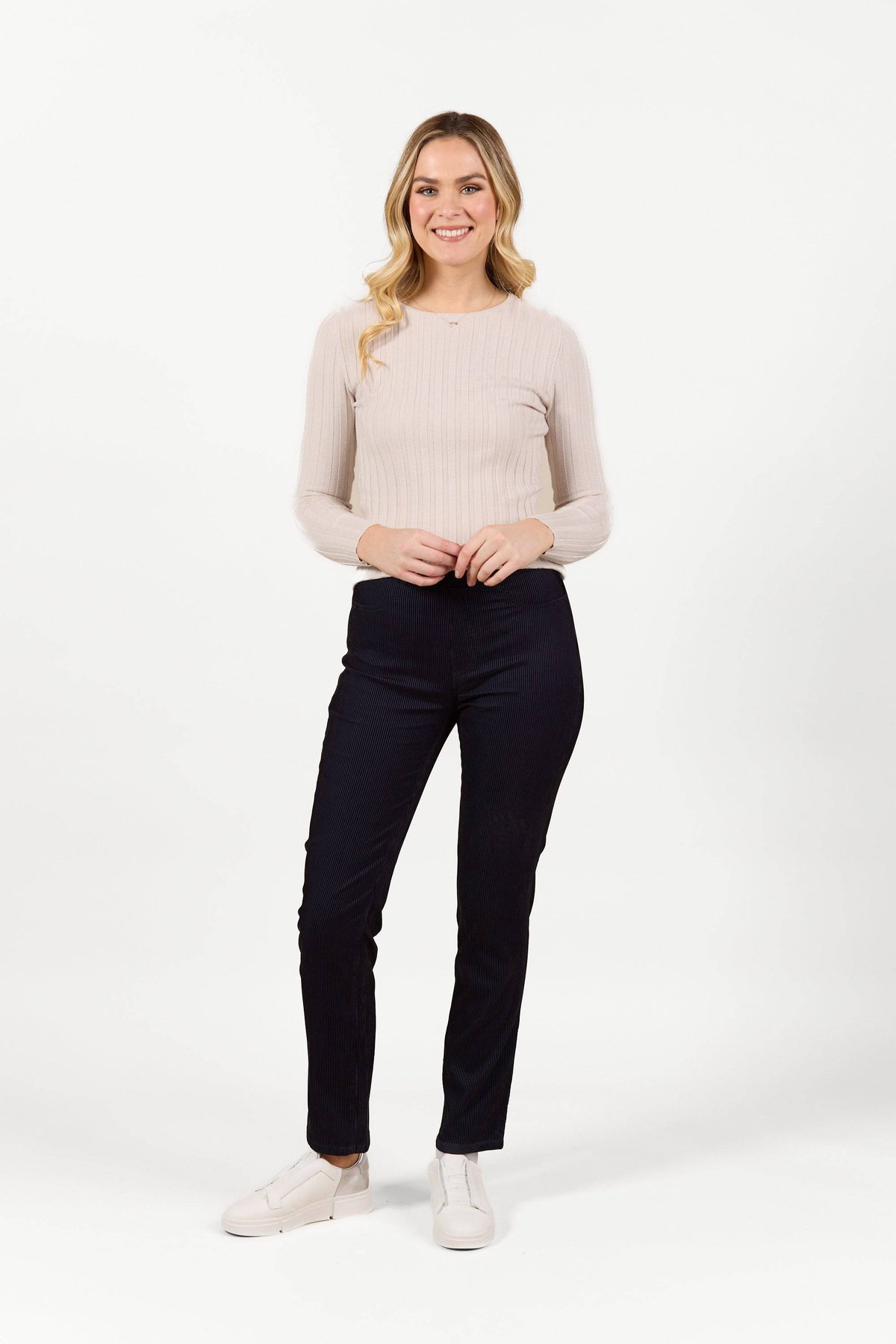 A woman with long blonde hair, wearing a light beige ribbed long-sleeve top, Bay Pull On - Navy Velvet pants by Vassalli, and white shoes, stands smiling against a plain white background.
