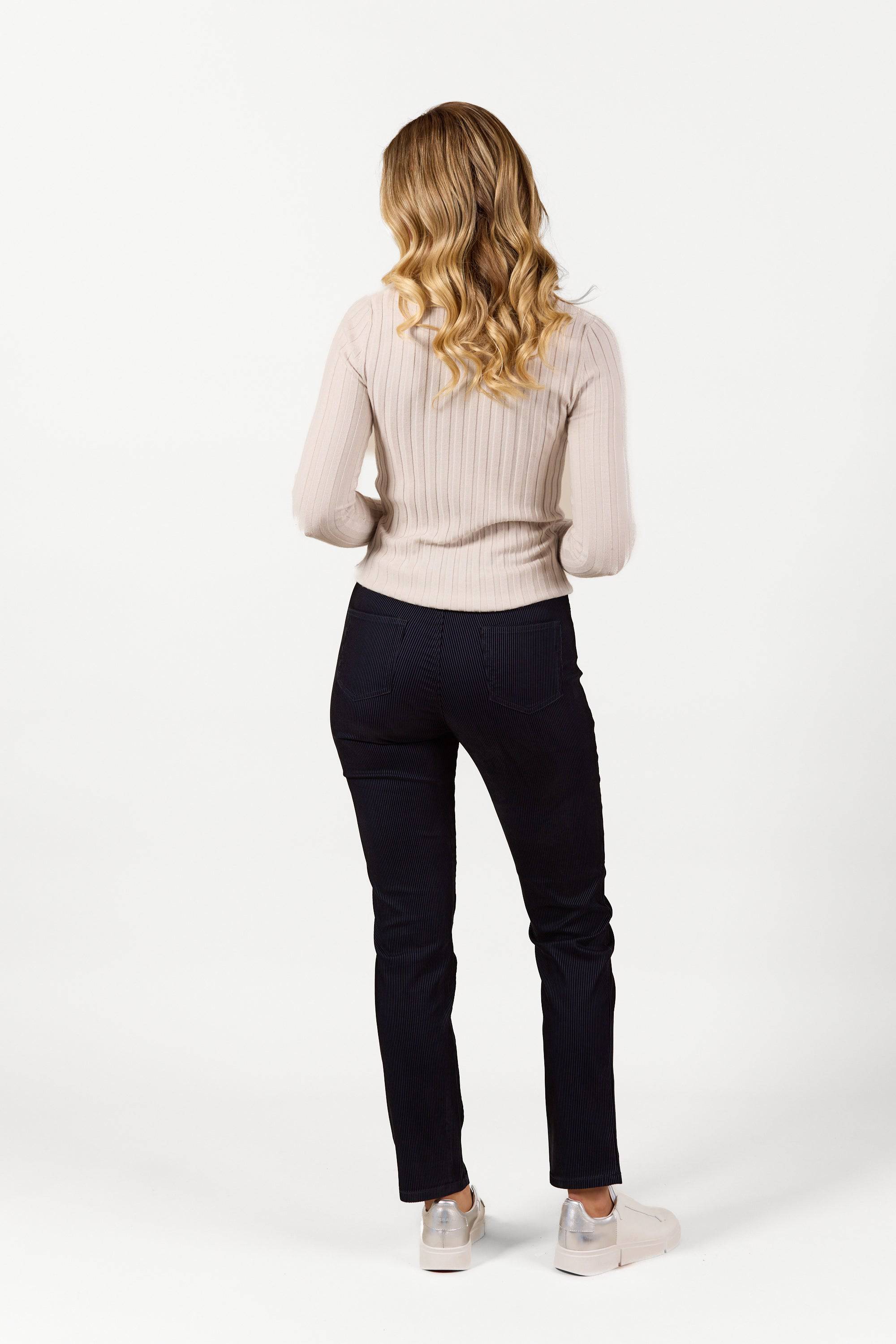 A woman with wavy blonde hair stands facing away, wearing a light ribbed sweater, white sneakers, and Vassalli's Bay Pull On pants in Navy Velvet against a plain white background.