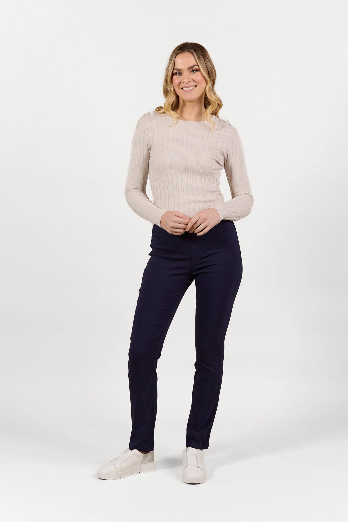 A woman stands smiling in Vassalli's Bay Pull On - Navy Bliss slim leg pants, a light beige ribbed long-sleeve top, and white slip-on shoes, posing against a plain white background.
