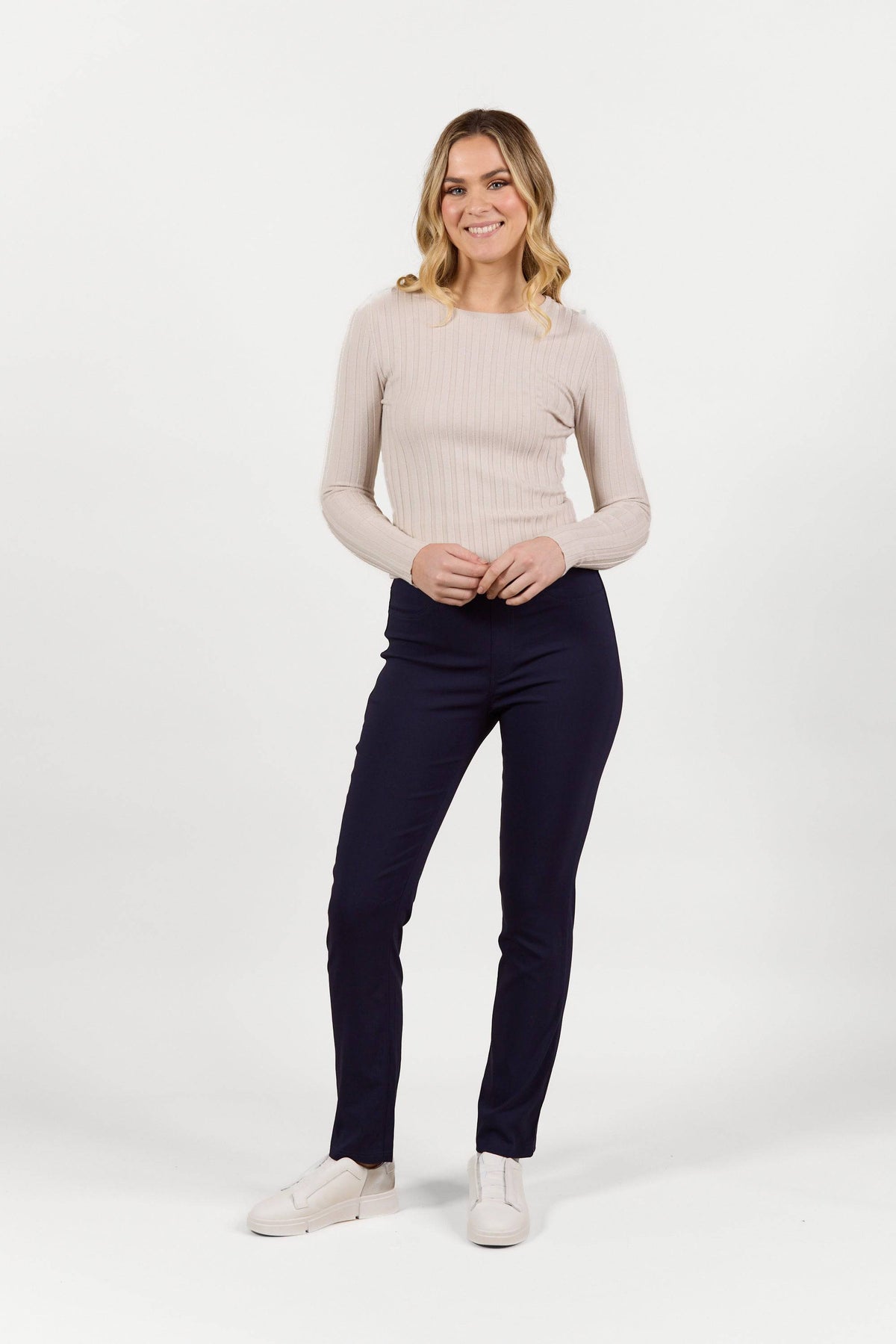A woman smiles against a white background, wearing a light beige ribbed long-sleeve top, white slip-on shoes, and Vassalli Bay Pull On - Navy Bliss slim leg pants with her hands loosely clasped in front of her.