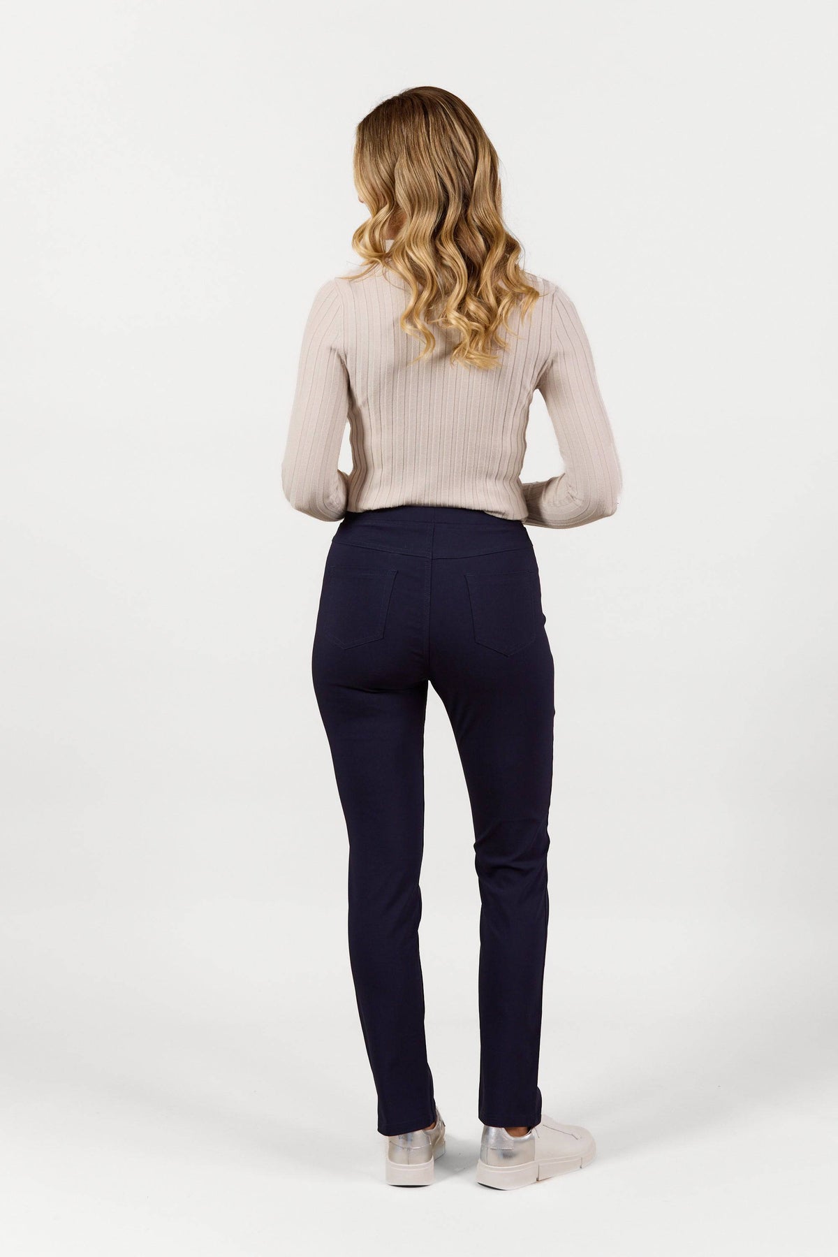 A person with long, wavy blonde hair stands facing away, wearing Vassalli's Bay Pull On - Navy Bliss slim leg pants, a light ribbed sweater, and light-colored shoes against a plain white background.
