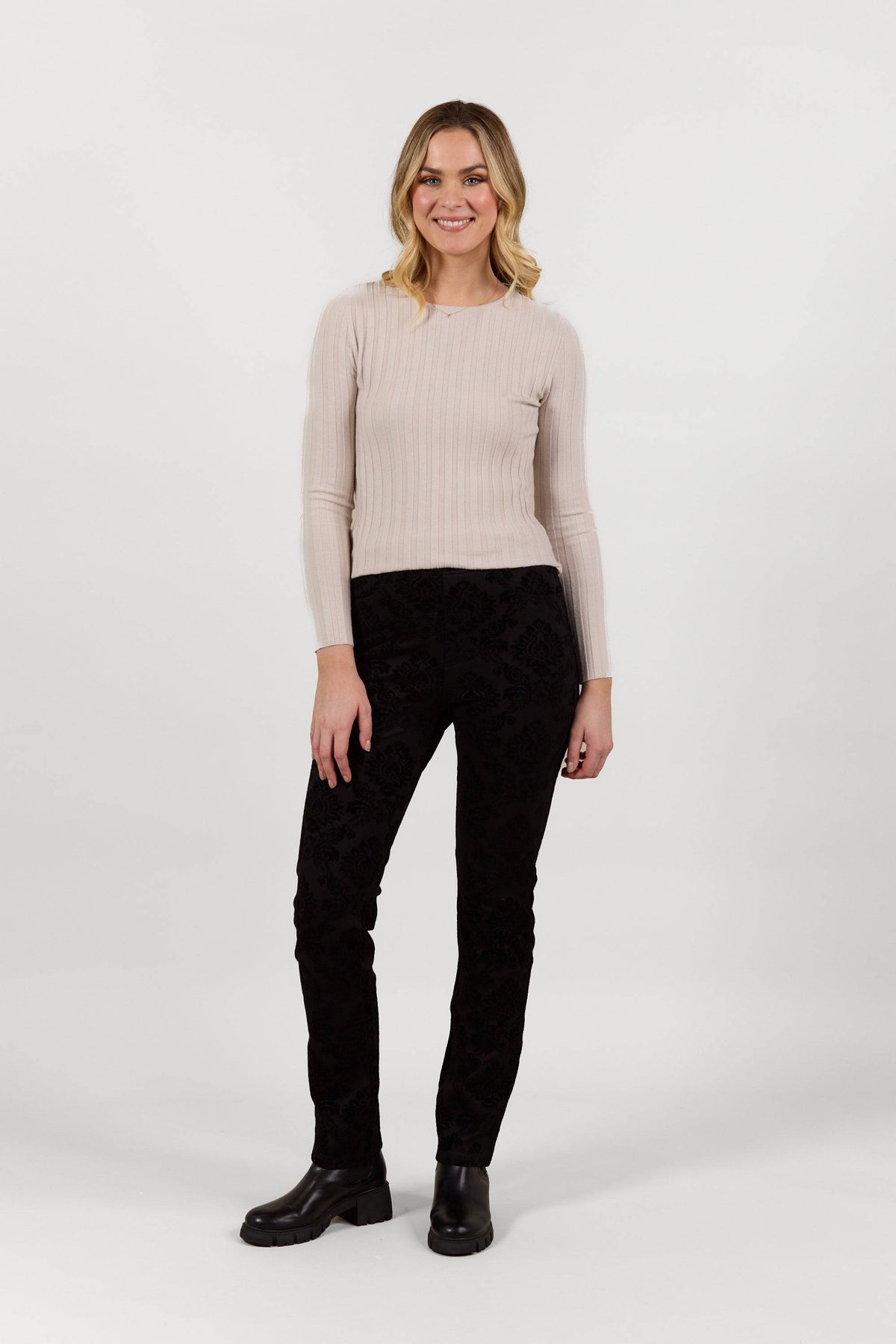 A woman smiles against a plain white background, wearing a light beige ribbed long-sleeve top and Vassalli Bay Pull On - Paris black slim leg pants in stretch fabric, paired with black boots.