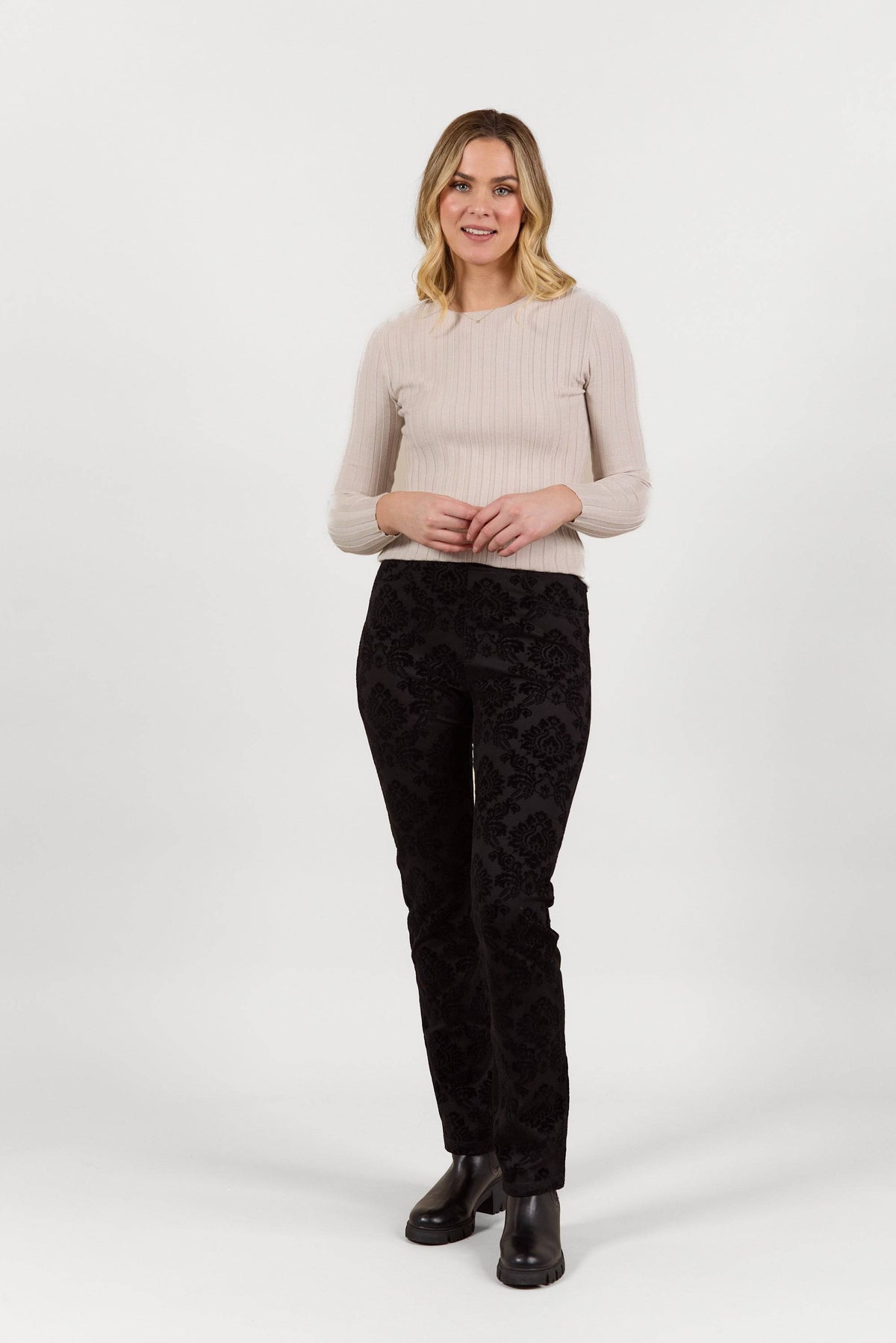A woman with blonde hair, wearing Vassalli's Bay Pull On - Paris black slim leg pants and a light beige long-sleeve top, stands against a white background, smiling with her hands loosely clasped.