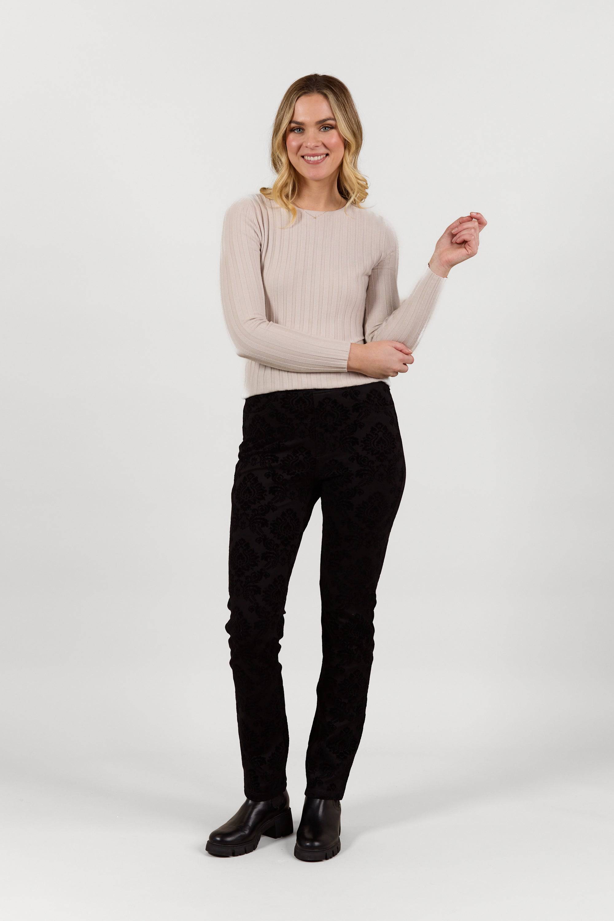 A woman with blonde hair, wearing Vassalli Bay Pull On - Paris dark slim leg pants, a light beige ribbed sweater, and black boots, stands smiling against a plain white background.