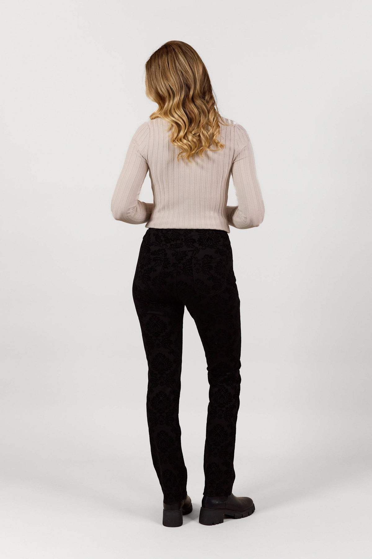 A woman with wavy blonde hair, facing away, wears Vassalli's Bay Pull On - Paris black slim leg pants and a light ribbed sweater with black shoes, set against a plain white background.