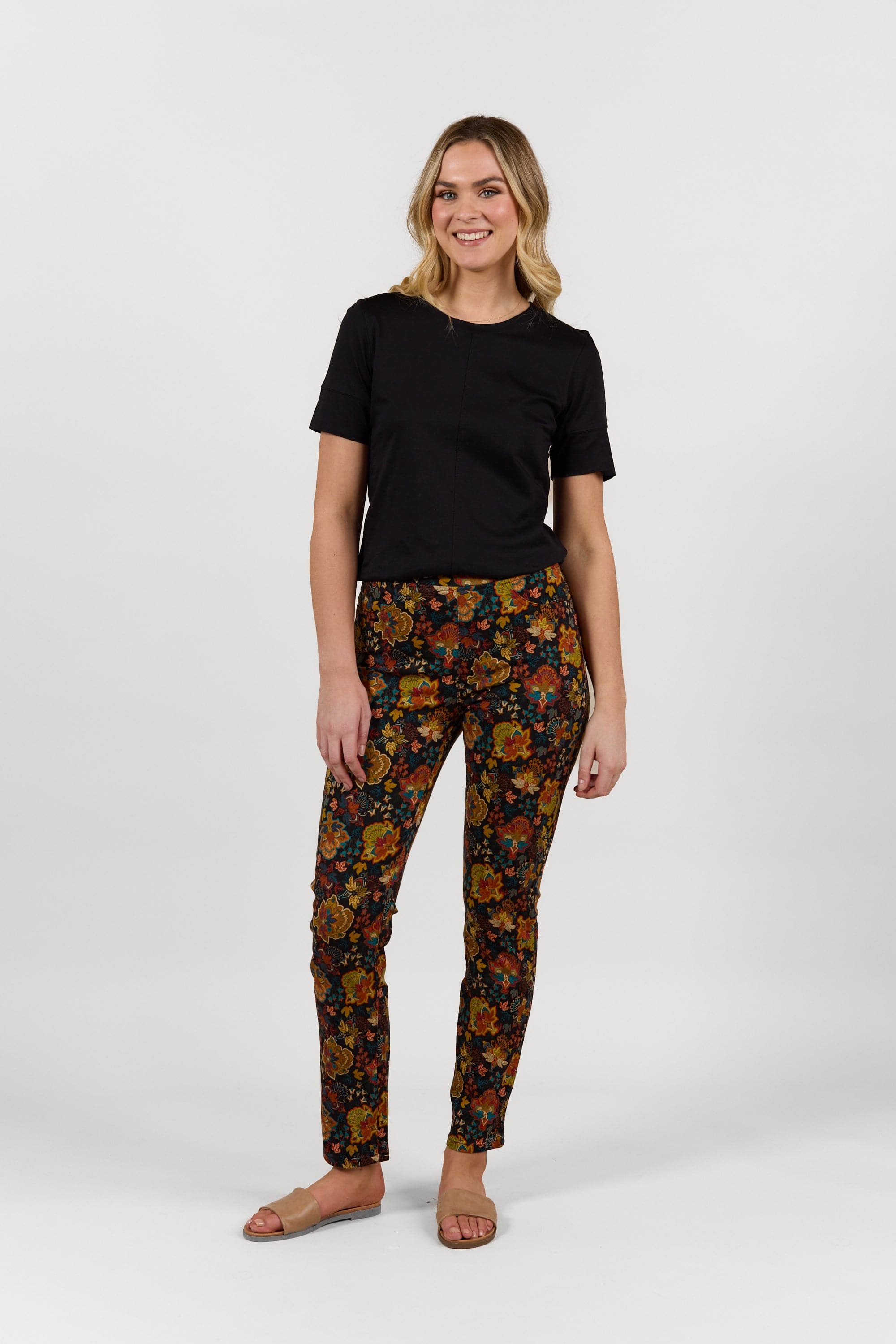 A woman stands smiling in the Vassalli Bay Pull On - Royal slim leg pants with an elastic waistband and orange-yellow floral print, paired with a black short-sleeved top and beige sandals against a plain white background.