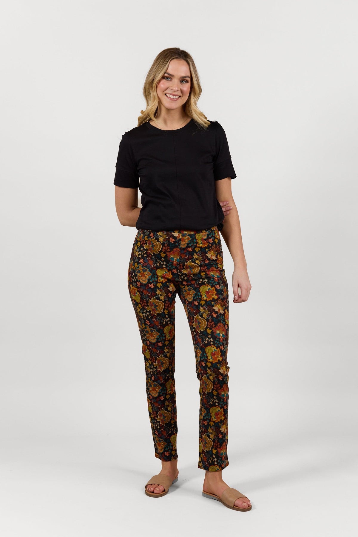 A woman with wavy blonde hair smiles against a white background, wearing a black t-shirt and Vassalli Bay Pull On - Royal floral slim leg pull-on pants with an elastic waistband, paired with tan sandals. One arm rests across her waist.