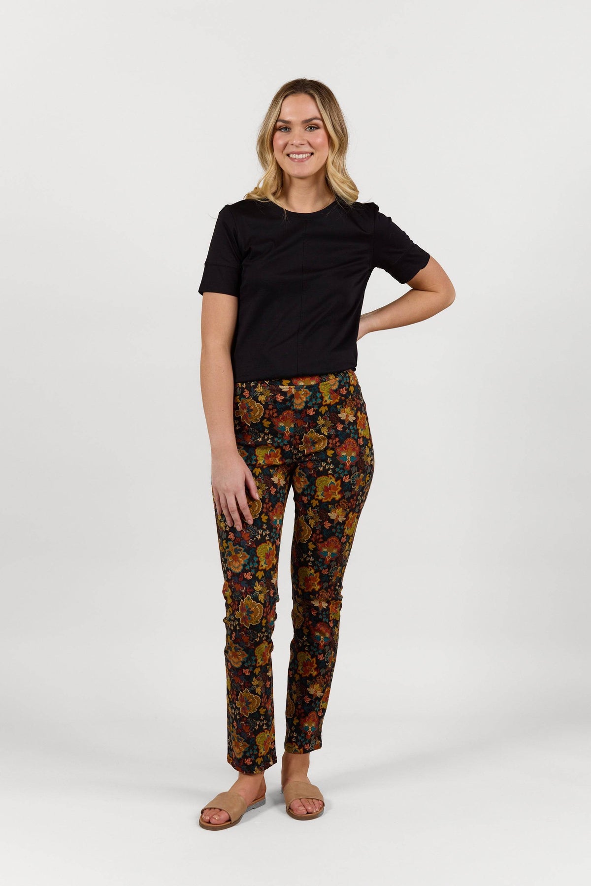 A woman smiles against a white background, wearing Vassalli's Bay Pull On - Royal slim leg pants with an elastic waistband, a black short-sleeve top, and tan sandals. One hand rests on her hip as she looks straight ahead.