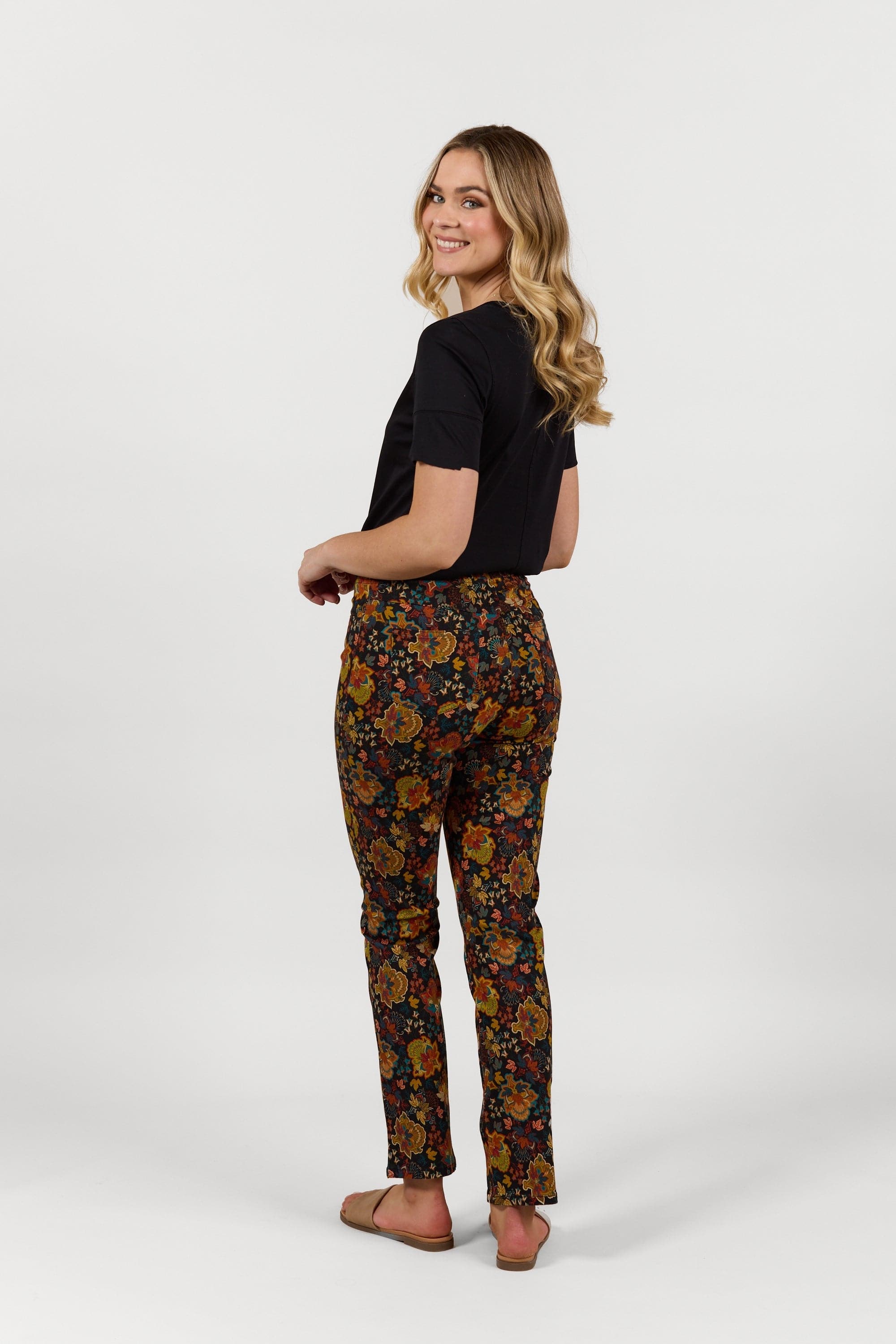 A woman with long blonde hair smiles over her shoulder, wearing the Vassalli Bay Pull On - Royal slim leg elastic waistband pants and a black short-sleeve top, posing against a plain white background in tan shoes.