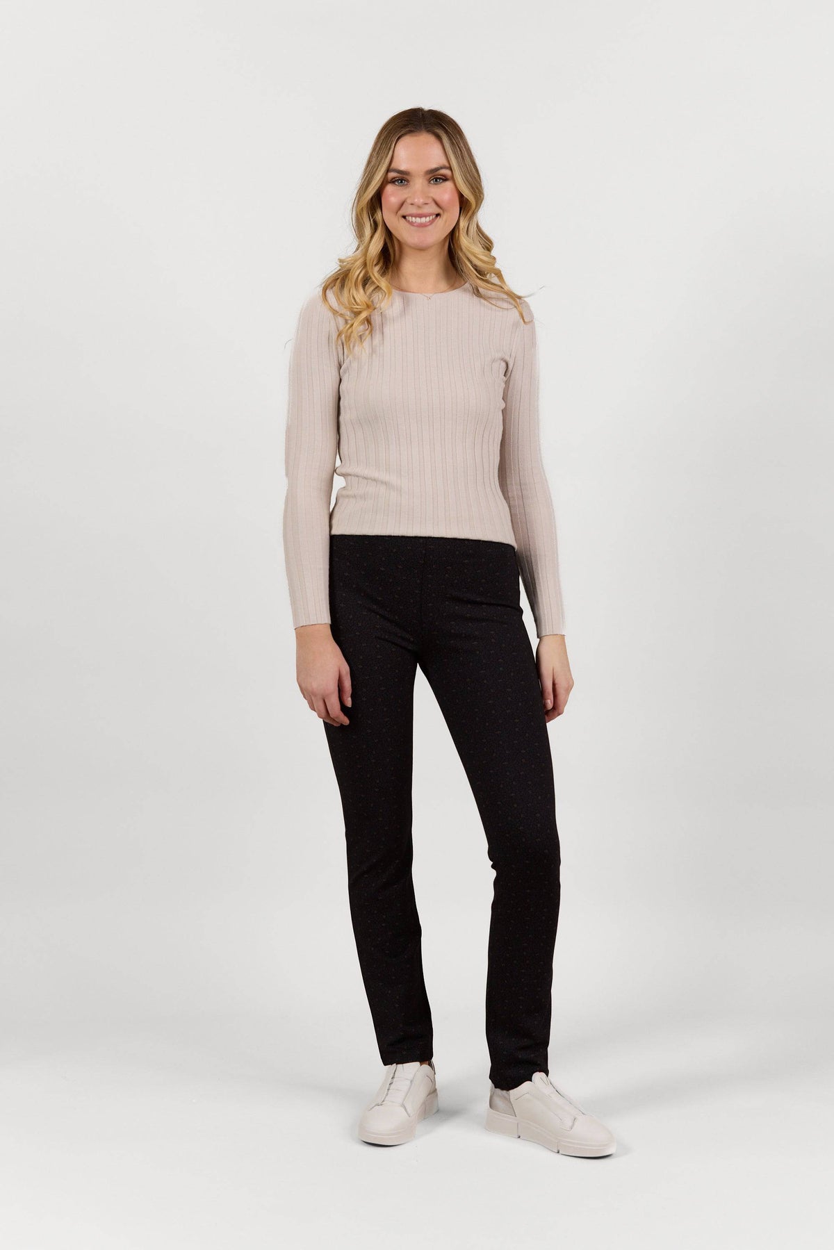 A woman with long blond hair smiles while wearing the Vassalli Bay Pull On - Sprig black slim leg pants, a light beige ribbed sweater, and white shoes, posing against a plain white background.