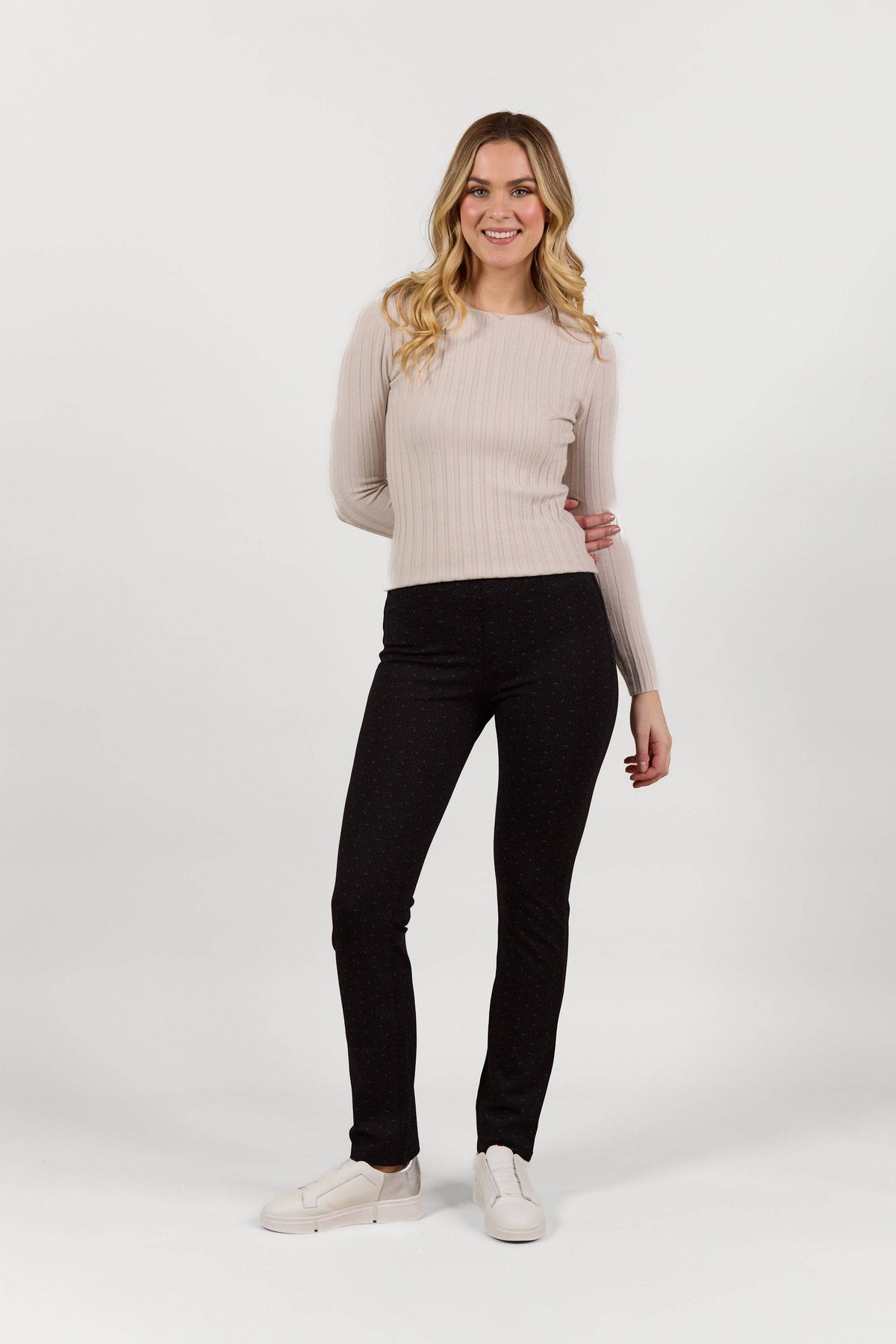 A woman with long blonde hair smiles against a plain white background. She wears a light beige ribbed sweater, dark slim-leg stretch pants, and Vassalli Bay Pull On - Sprig slip-on shoes.